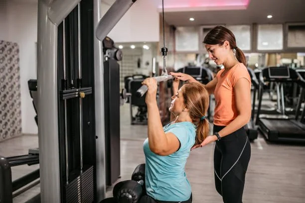 Two women at a gym, one using a cable machine for strength training and the other assisting or instructing.