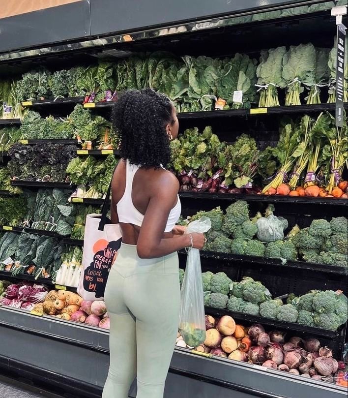 A woman shopping for vegetables in a grocery store, wearing a white sports bra and light green leggings, carrying a tote bag and a plastic bag of produce, standing in front of a vegetable display with leafy greens, broccoli, and root vegetables.