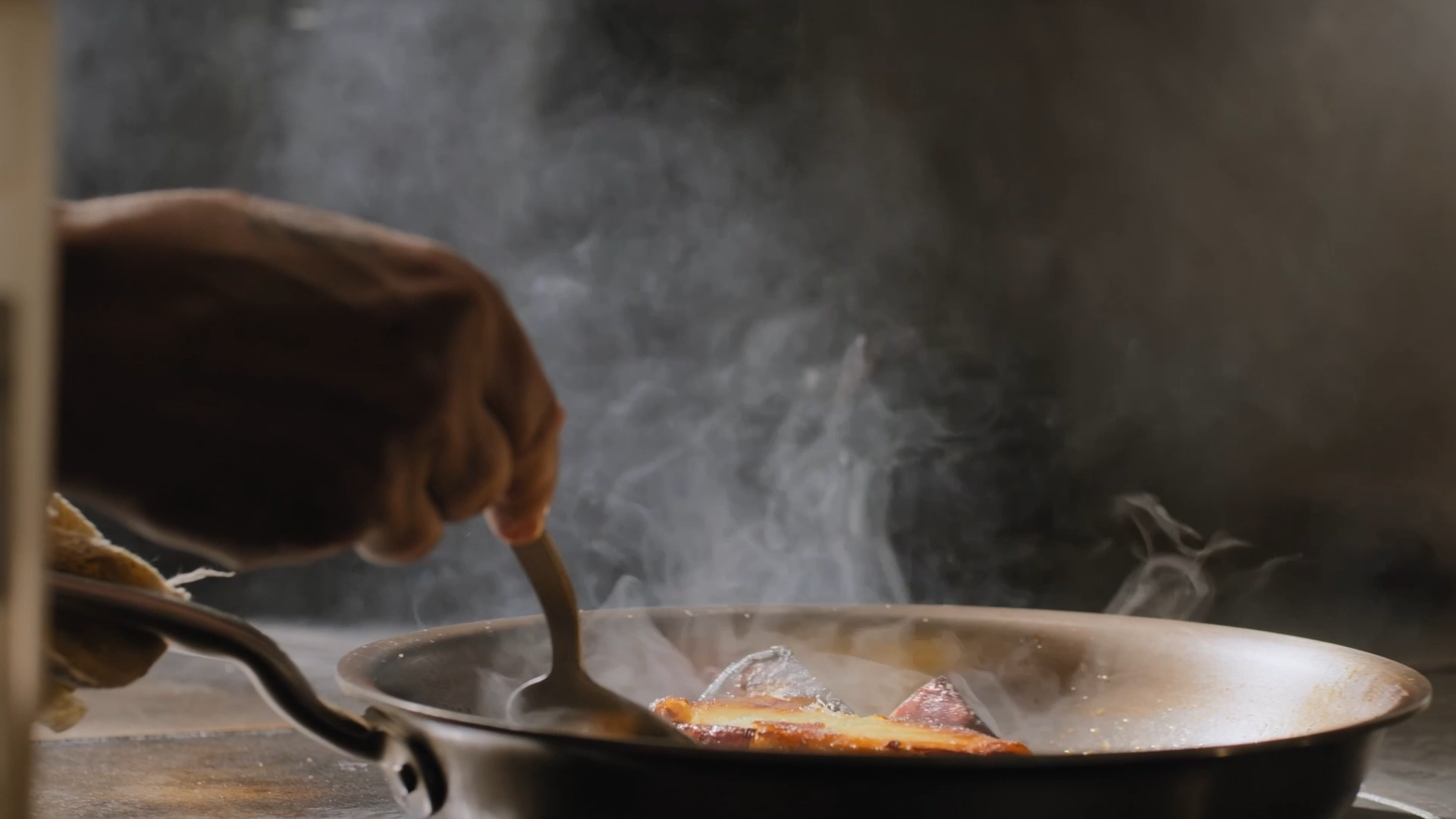 A person wearing a brown glove uses a spoon to cook food in a steaming pan, with smoke rising in the kitchen.