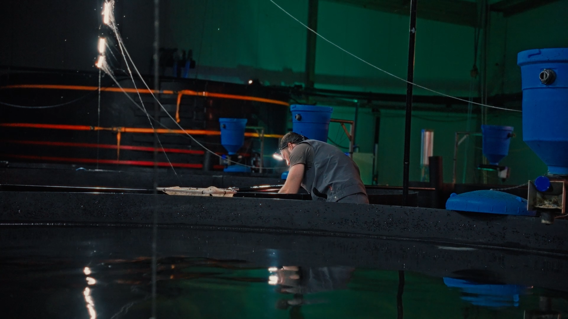 A person working with equipment in an industrial or manufacturing setting, surrounded by blue containers and machinery, with reflections visible on a water surface in the foreground.