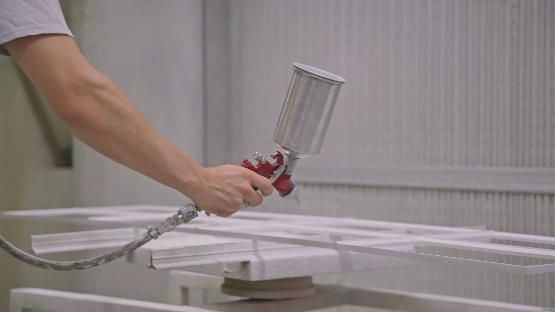 Person spraying white frames with a paint spray gun in a workshop or manufacturing setting.