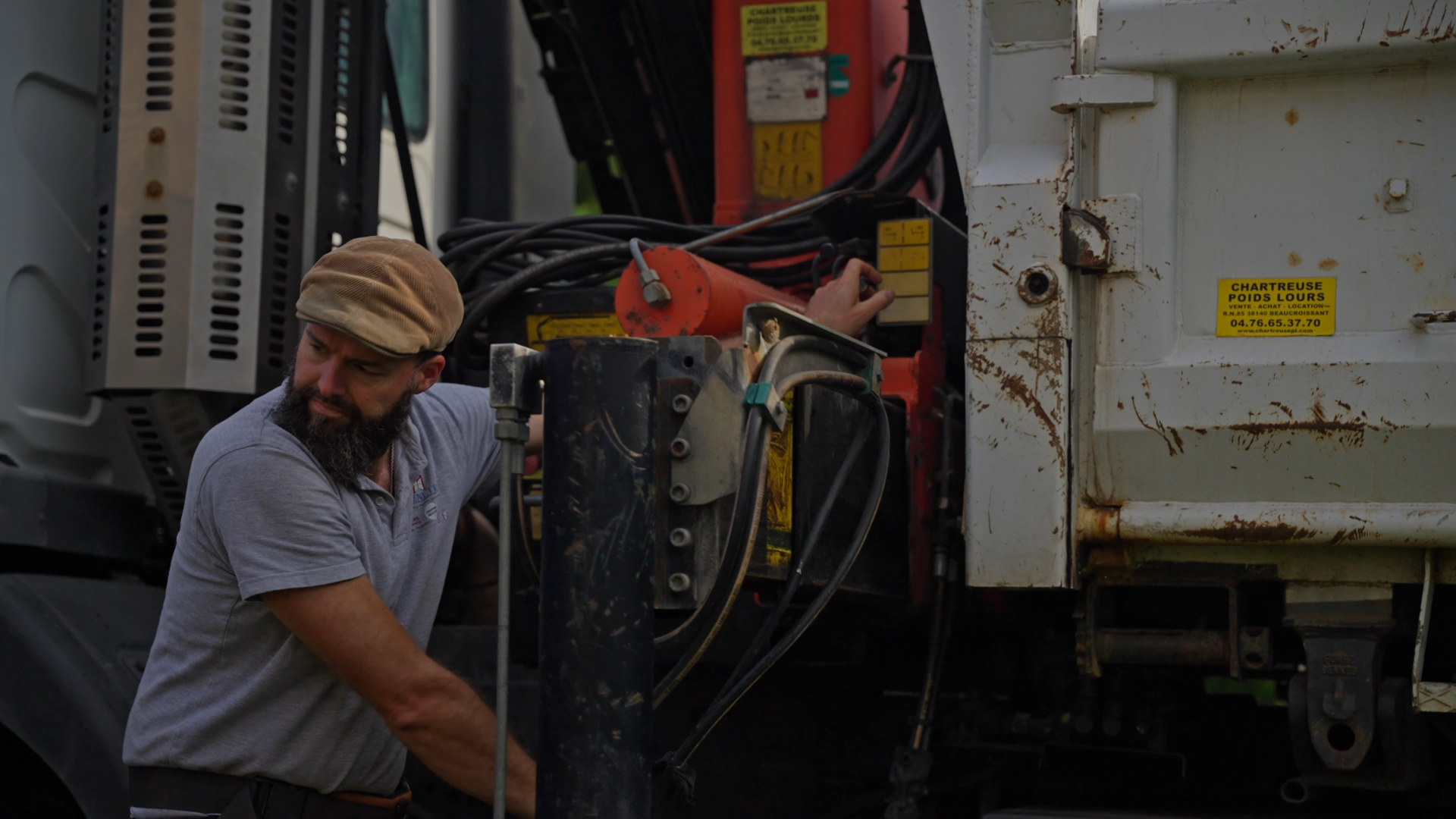 A man wearing a tan cap and gray polo shirt working on a large piece of machinery, adjusting electrical connections on a construction or industrial vehicle.