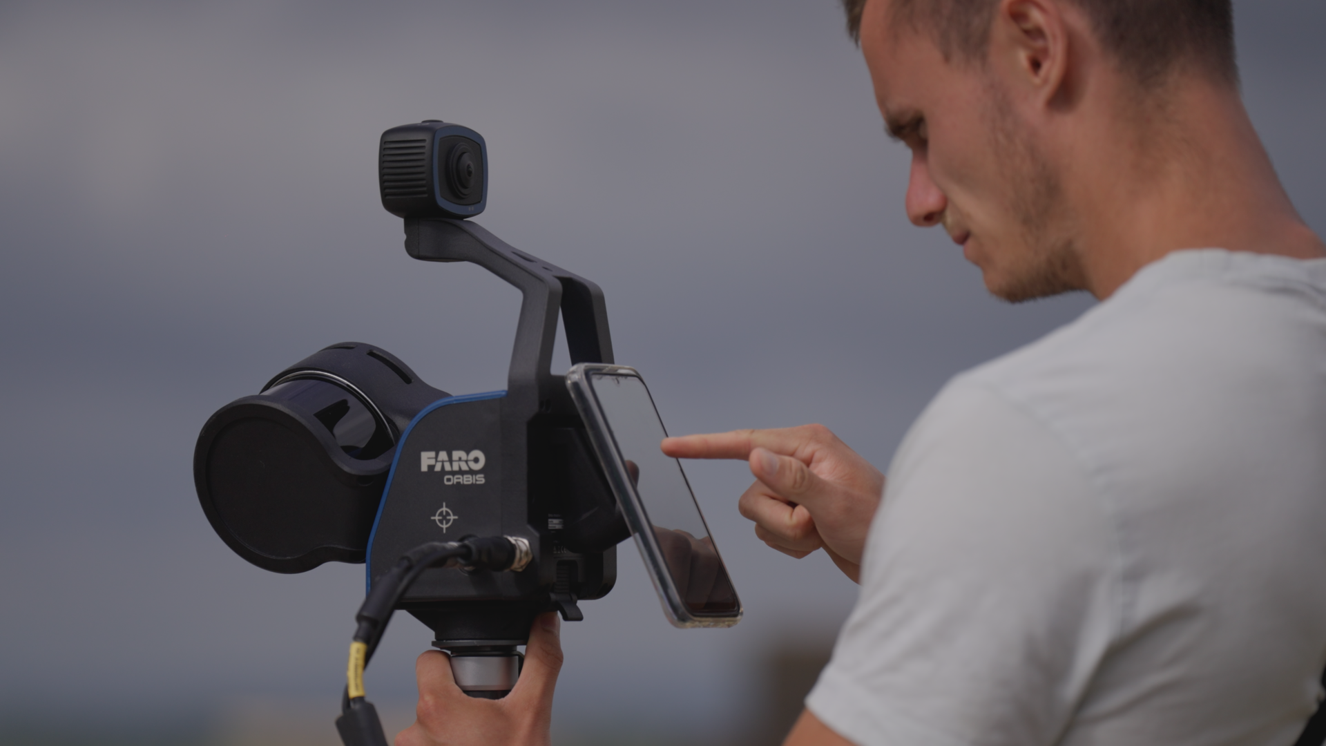 A man pointing at his smartphone screen while operating a drone controller with a mounted camera, outdoors during cloudy weather.