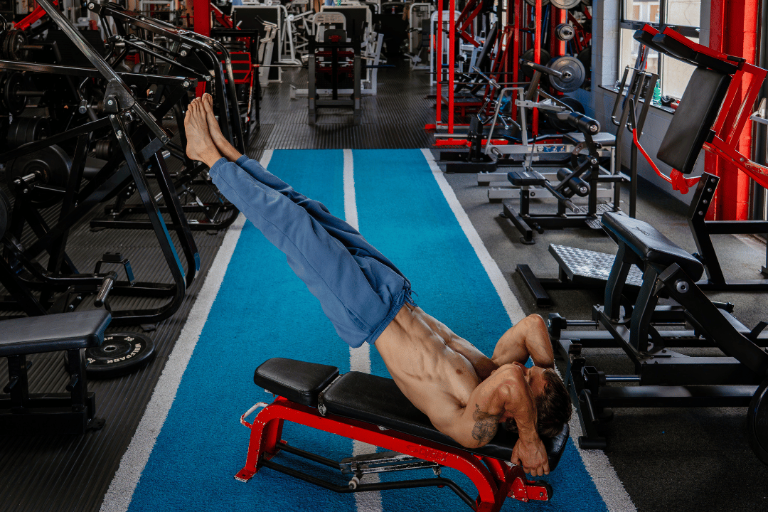 A shirtless man with tattoos do a leg raise exercise on a workout bench in a gym with various fitness equipment around. reps gym omagh