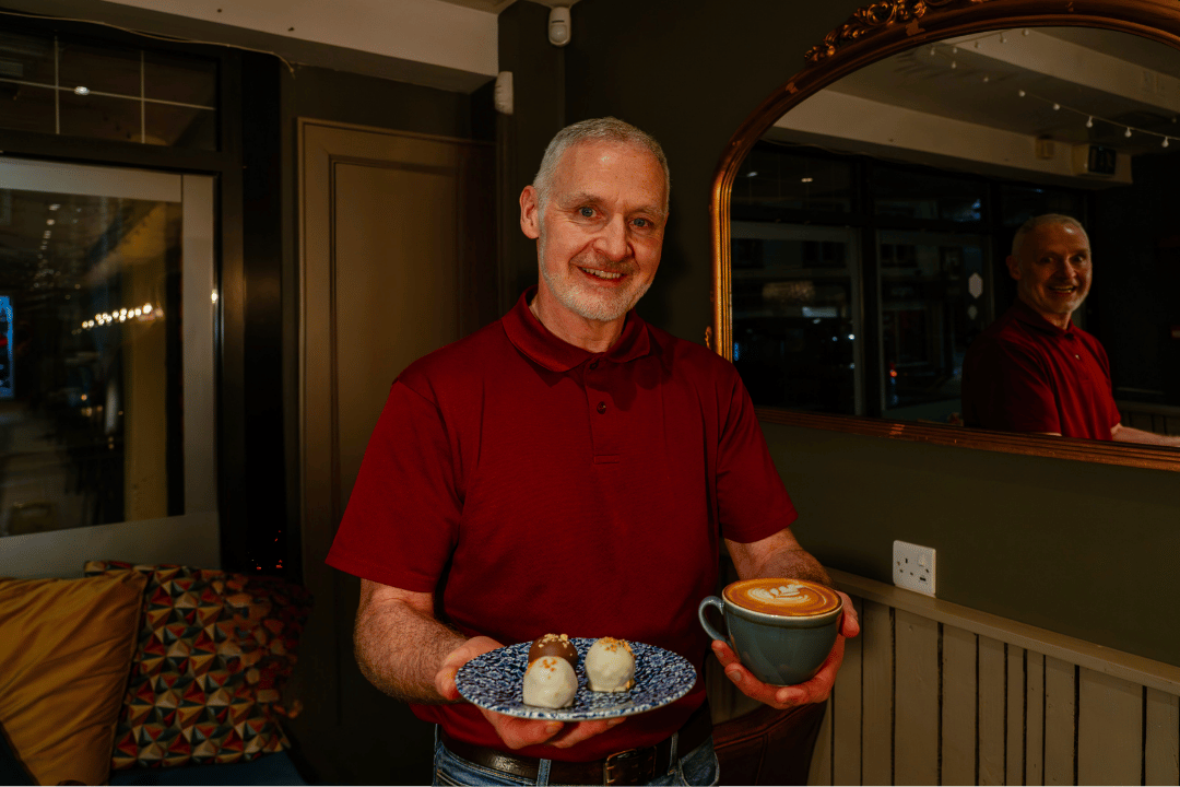 A man with gray hair and a beard smiling while holding a plate with two desserts and a cup of coffee inside a café at night. Sandwich co omagh