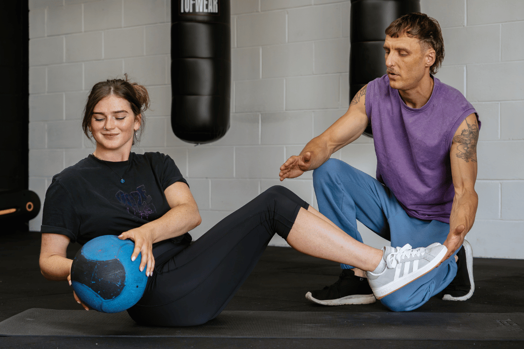 A woman sitting on a gym mat, holding a blue medicine ball with her hand, while a trainer assists her in performing a leg exercise with her foot, inside a gym with hanging punching bags. Independent gym in Northern Ireland featured in reps gym