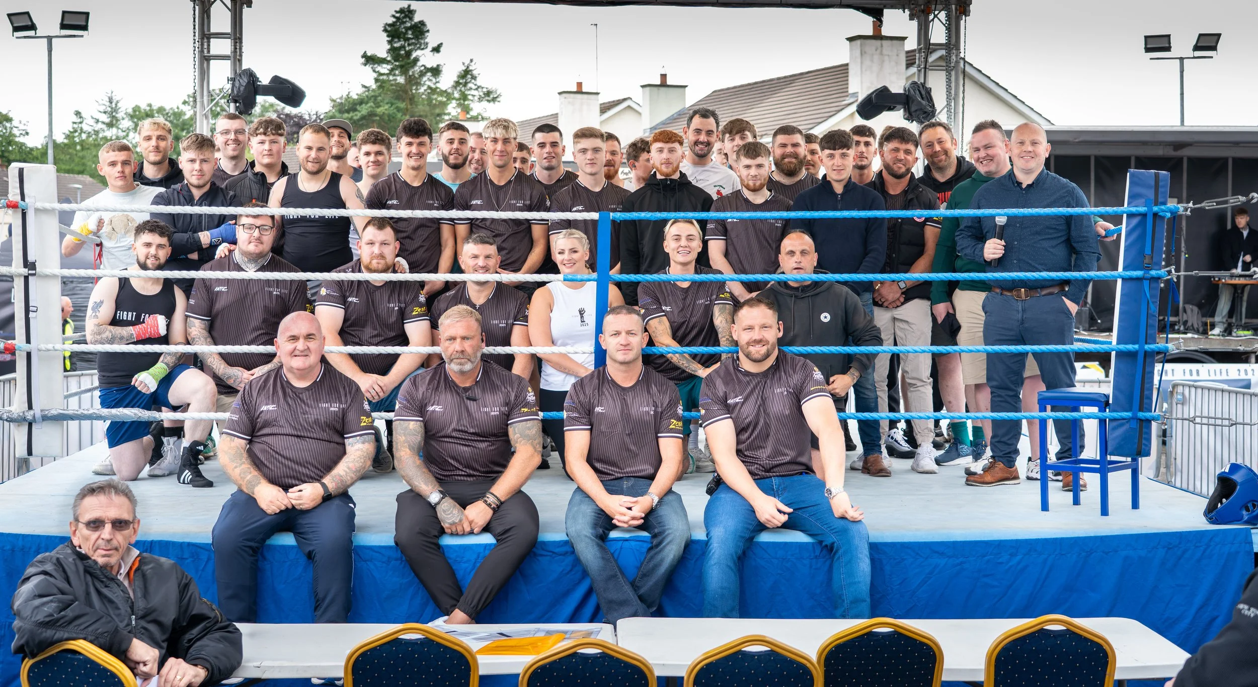 Group of people, including boxers and event staff, gathered in a boxing ring outdoors during daytime, with some smiling and others posing for the photo. after a charity boxing event in omagh called fight for life
