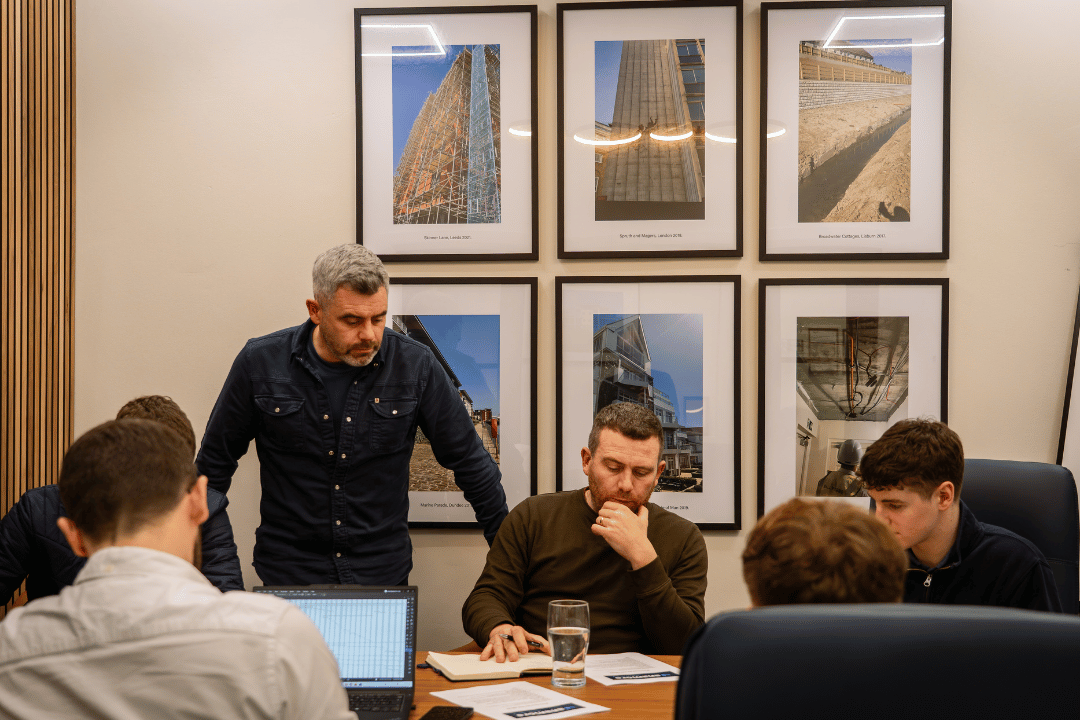A group of five men in a meeting room, with framed architectural photos on the wall behind them. One man is standing, while the others are seated, engaged in discussion, with one taking notes and others working on laptops.