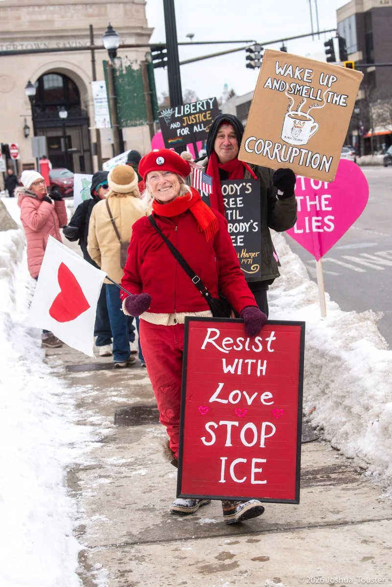 Protesters marching on a snow-covered sidewalk holding signs  including a woman with a sign that says 'Resist with Love, Stop Ice' and a man holding a sign that says 'Wake Up and Smell the Corruption'.