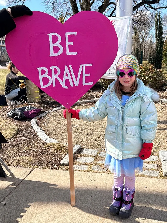 A young girl wearing sunglasses, a rainbow beanie standing outdoors, holding a large pink heart-shaped sign that says 'BE BRAVE'.