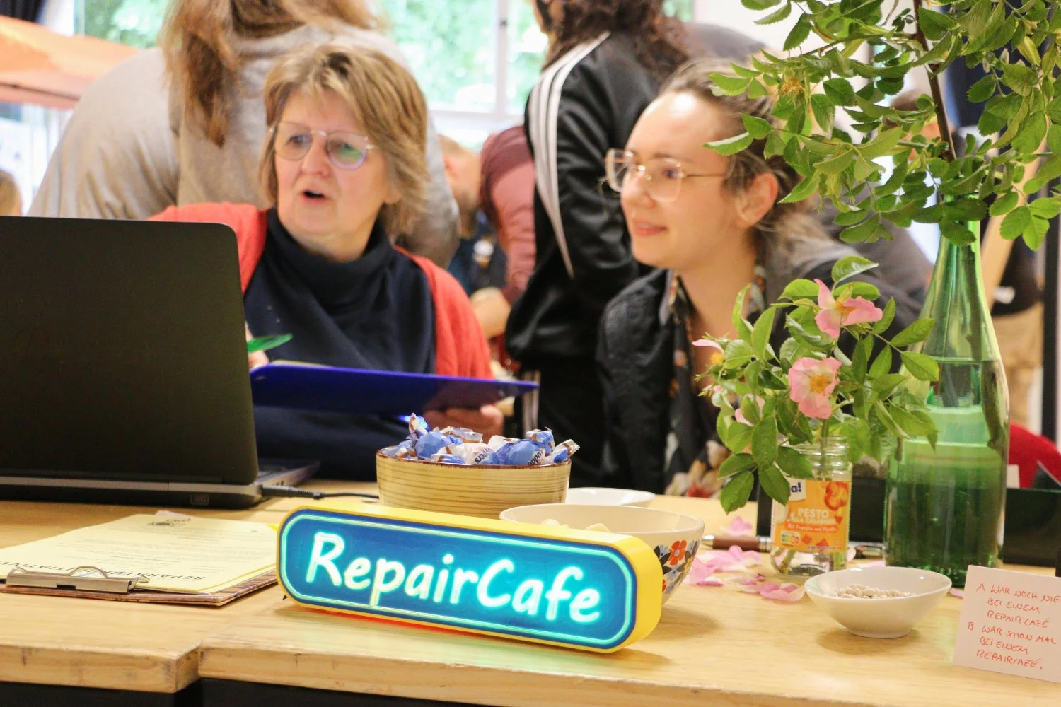 Two people looking at a laptop with a led sign "repair café" in front of them