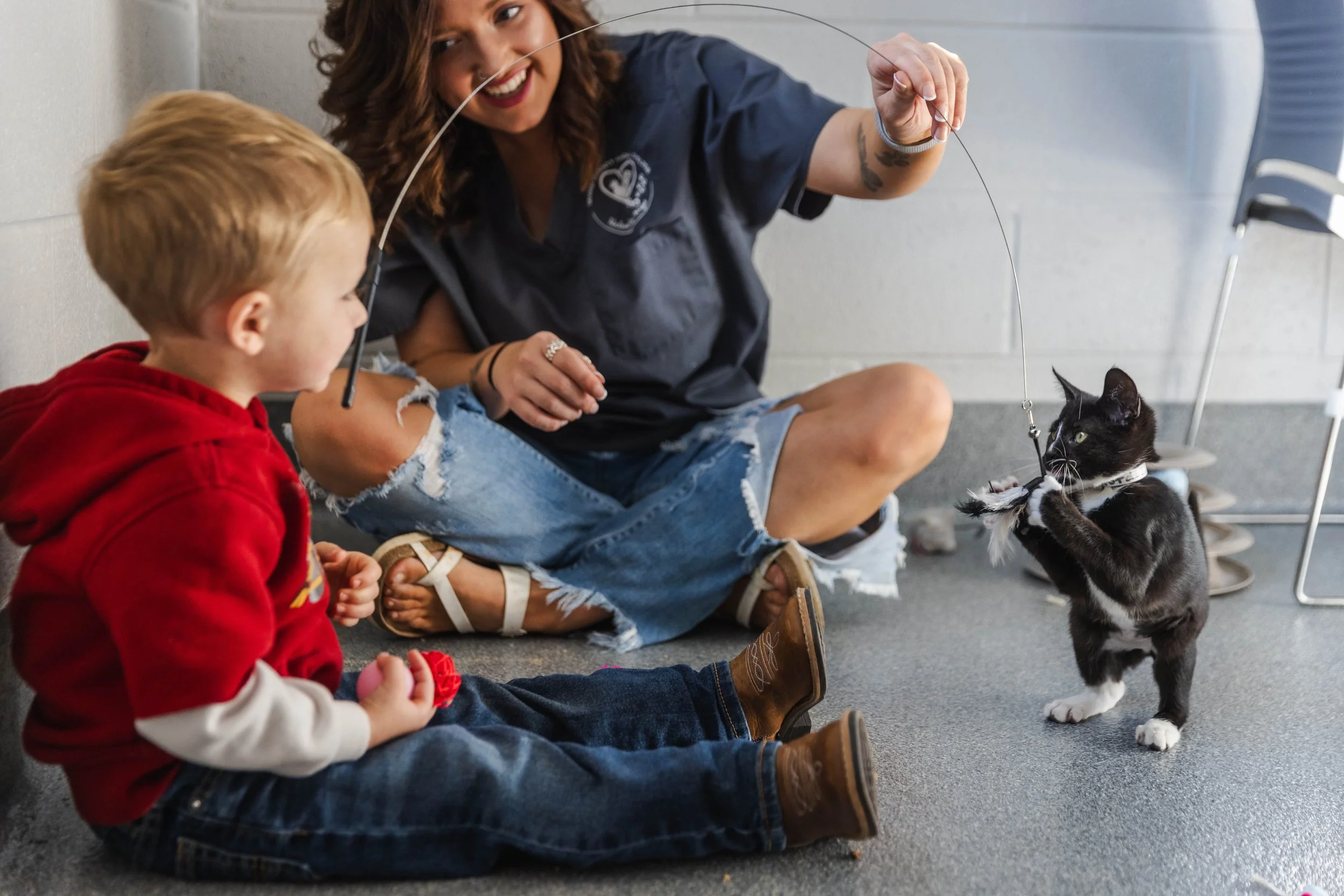 A little boy in a red jacket is playing with his mom and a kitten with a string at Sevier County Animal Shelter.