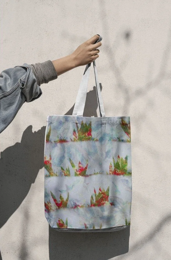 Hand hält eine bunte Stofftasche an einem schwarzen Haken an einer weißen Wand, Sonnenlicht wirft einen Schatten.