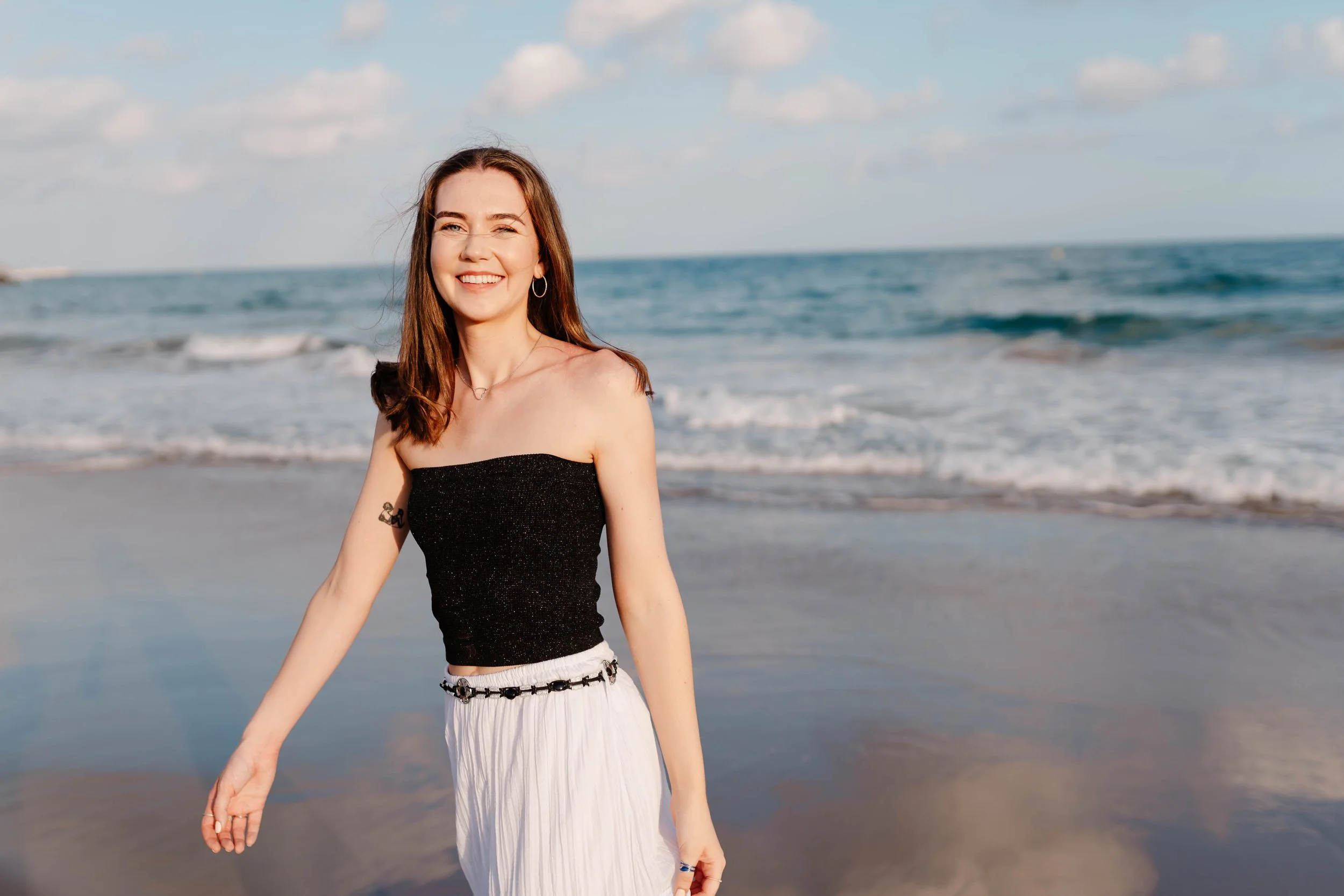 A woman with long brown hair wearing a black strapless top and white pants standing on a beach with ocean waves behind her, smiling.