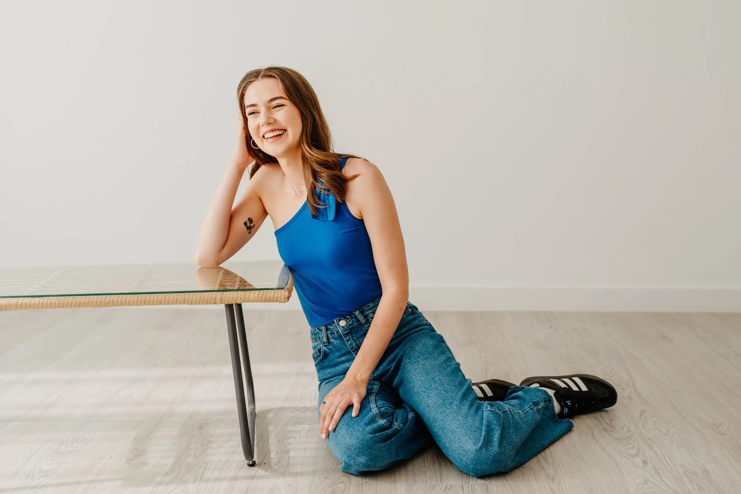 A young woman with brown hair, smiling and sitting on her knees, leaning against a glass-topped table with metal legs, wearing a blue tank top, baggy jeans, and black sneakers with white stripes, in a minimalist room with a light wooden floor and plain white wall.