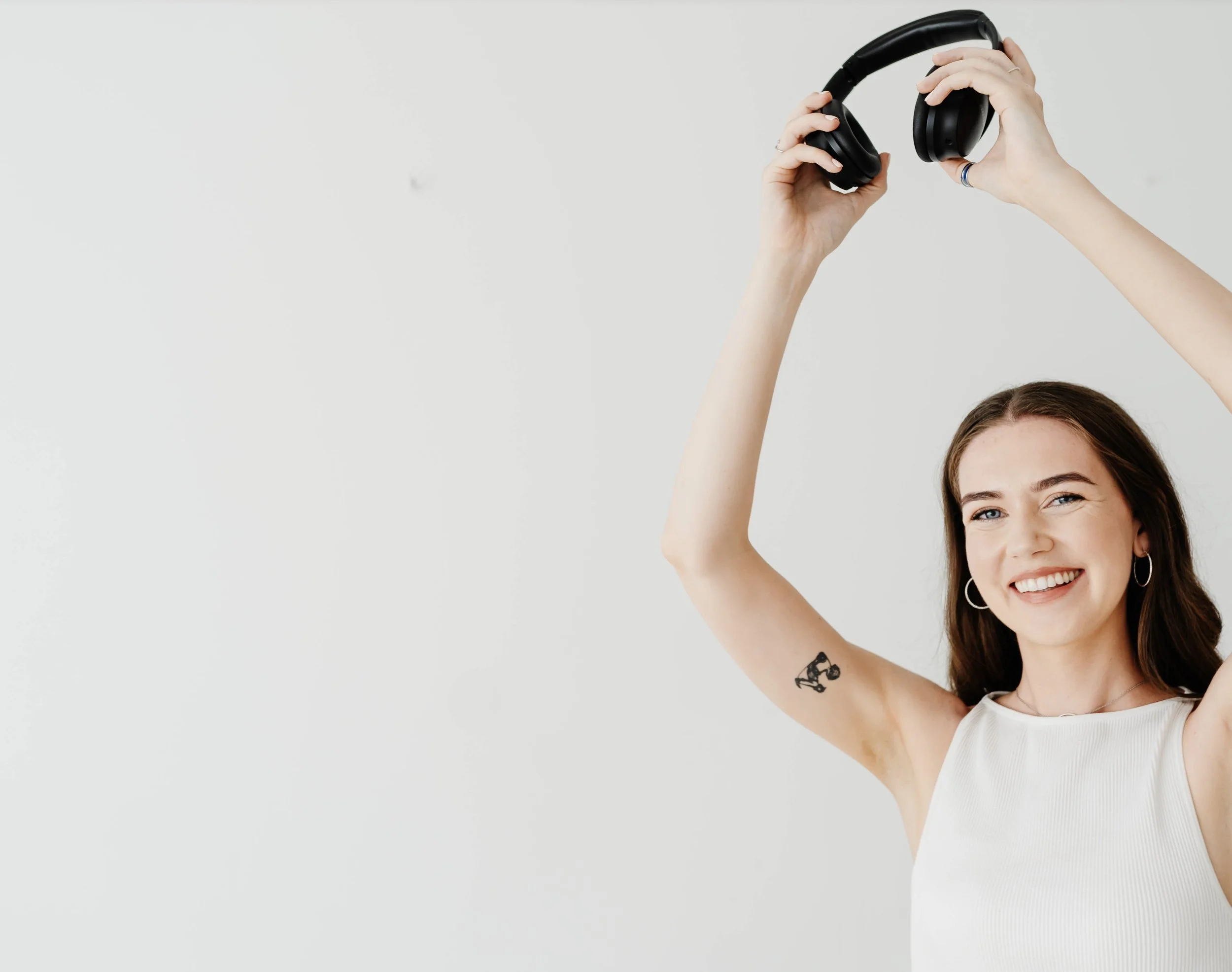 A young woman with long brown hair, earrings, and a small tattoo on her upper arm, smiling and holding headphones above her head against a plain white background.
