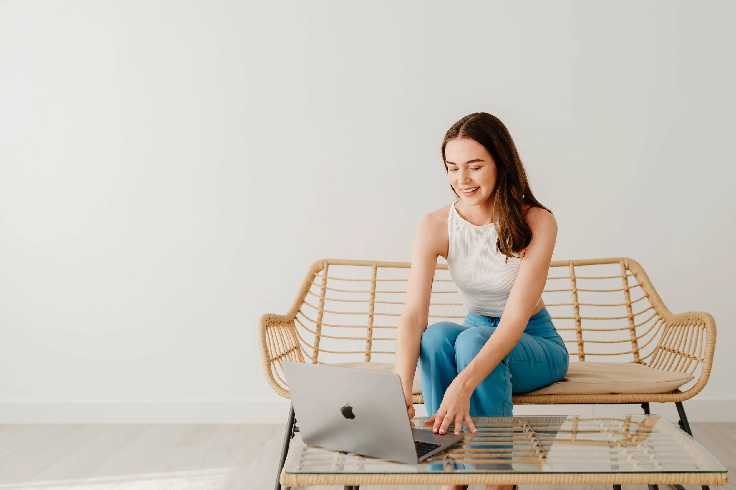 A woman in a white sleeveless top and blue pants sitting on a rattan bench with a laptop on a glass table in front of her, smiling and looking at the laptop.
