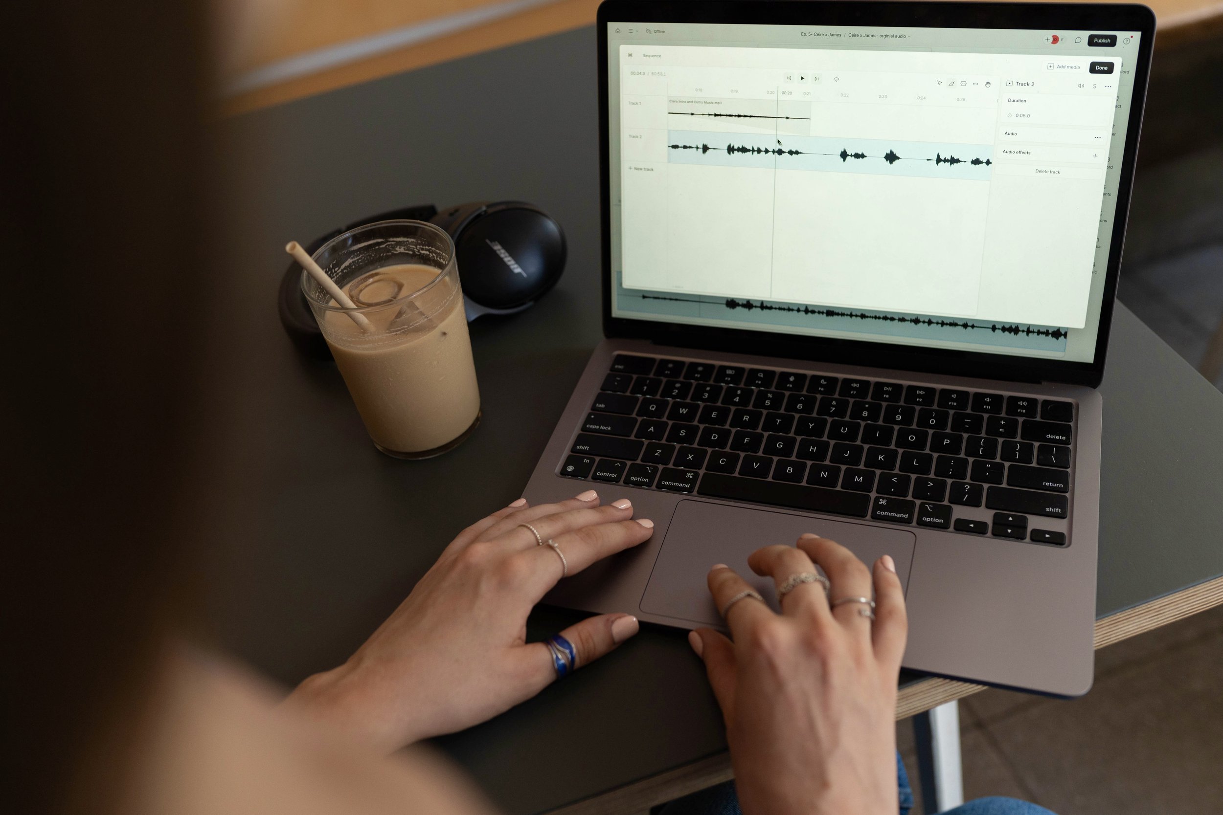 A person using a laptop with a digital audio editing program open, sitting at a table with a glass of iced coffee, a black wireless speaker, and a pair of black headphones nearby.