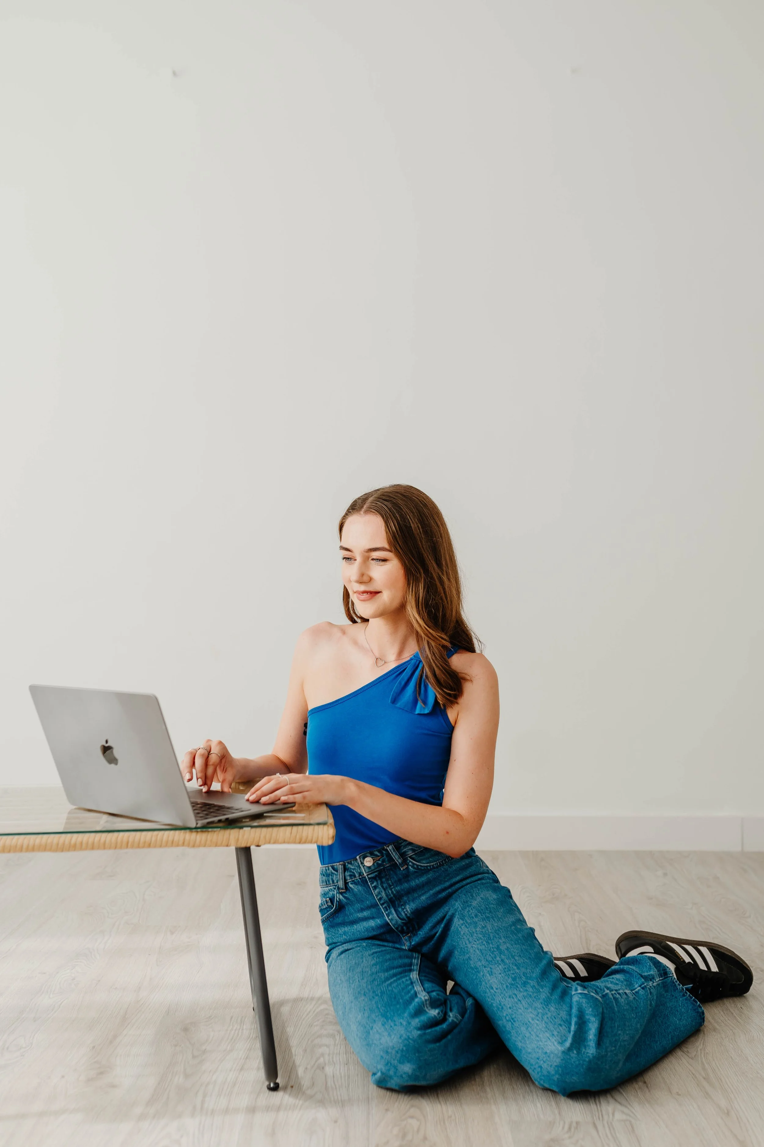 A young woman with long brown hair kneeling on a light wood floor, wearing a blue one-shoulder top and blue jeans, working on a silver laptop at a small wooden table in a plain white room.