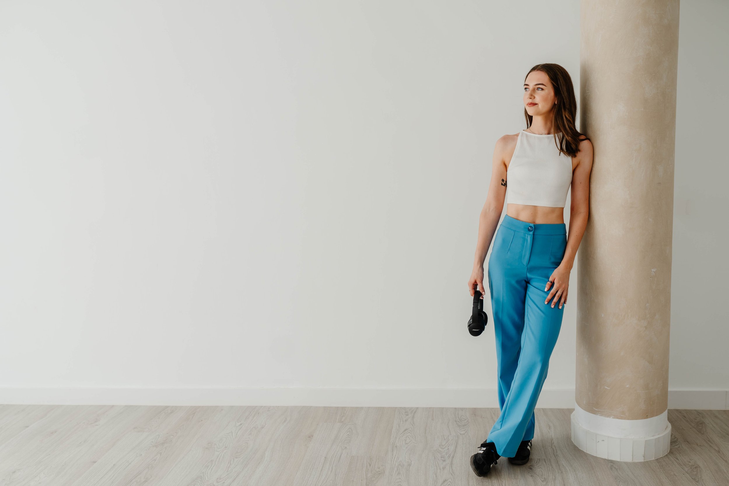 A young woman with brown hair leaning against a beige pillar, holding a small black camera, wearing a white crop top and bright blue pants, standing on a light wood floor in a minimalist room.