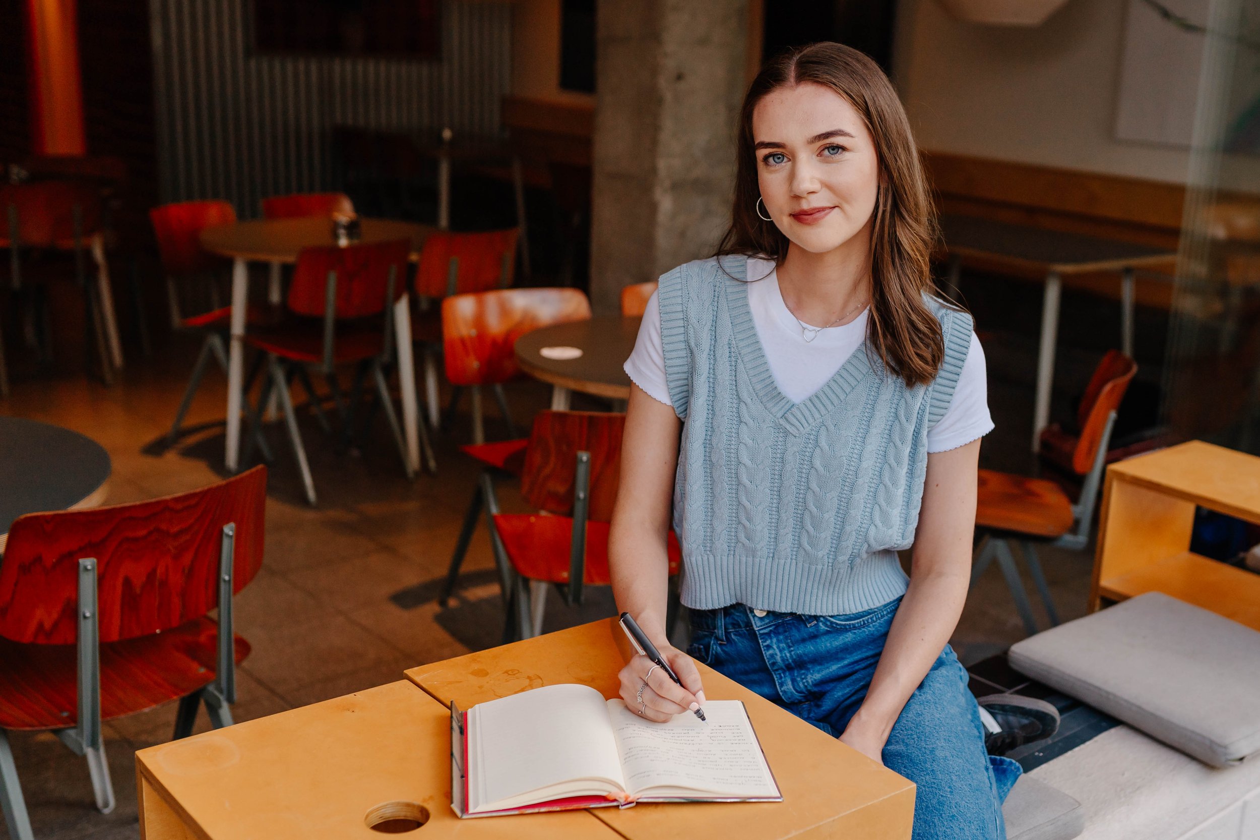 A young woman with brown hair, blue eyes, and hoop earrings sitting at a wooden table in a cozy cafe, holding a pen over an open notebook.