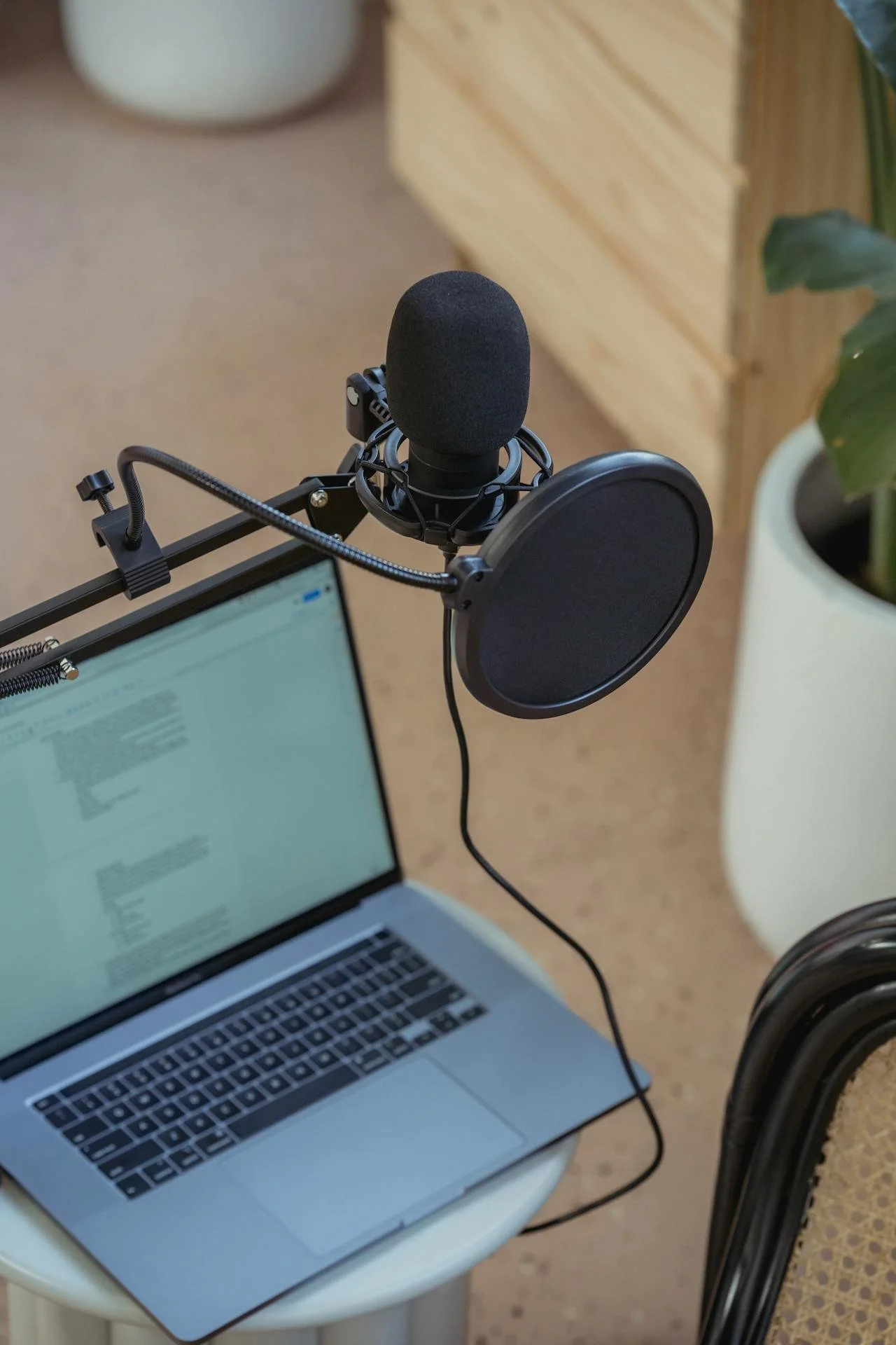 A podcasting setup on a small circular table includes a silver laptop with an open document, a black microphone with a pop filter mounted on a stand, and a laptop arm. In the background, there is a white cylindrical pot with a green plant and a wooden furniture piece.