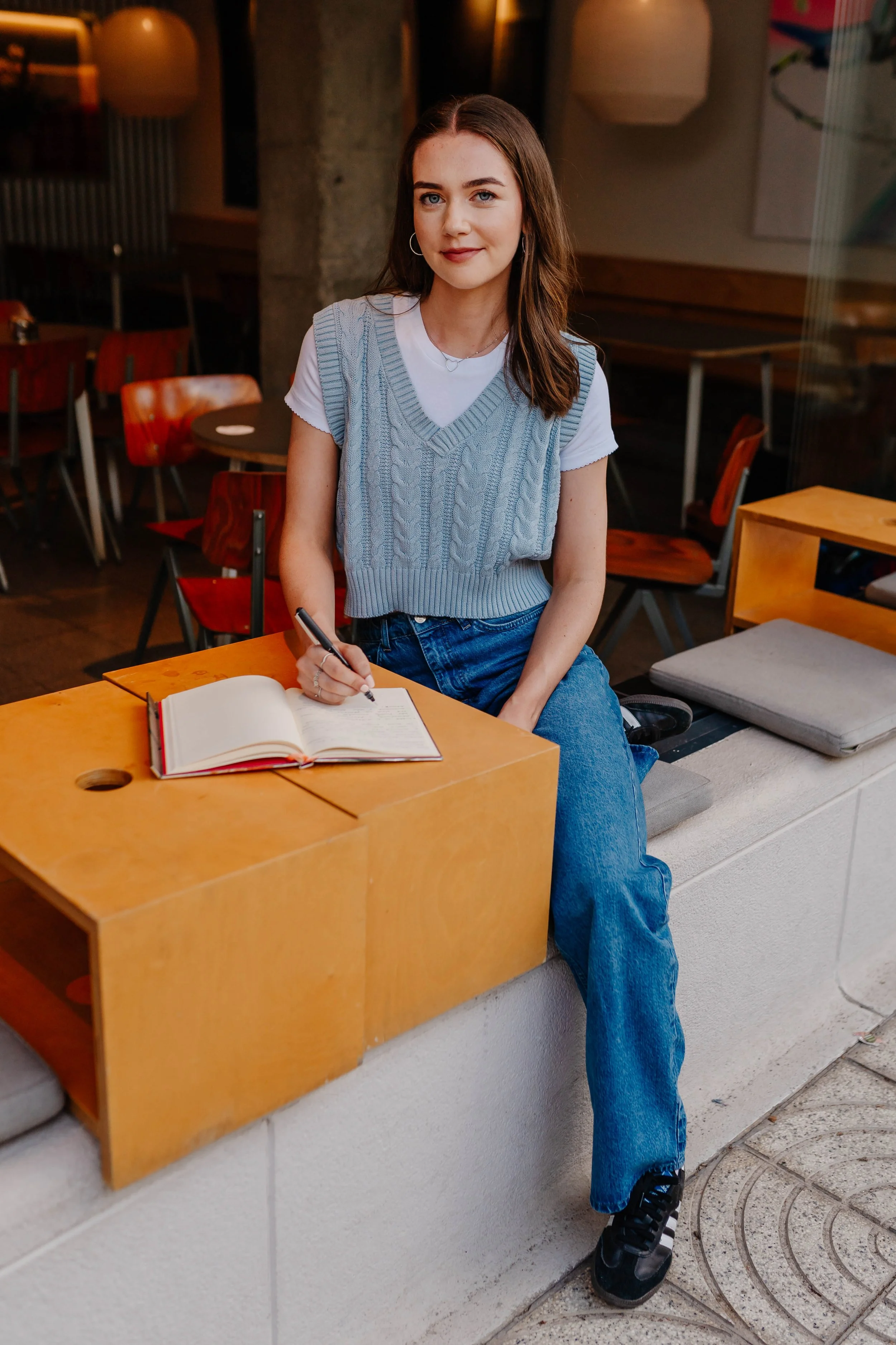 A young woman with brown hair, wearing a gray knit vest over a white T-shirt, sitting on a concrete ledge inside a cozy cafe, writing in a notebook with a black pen.