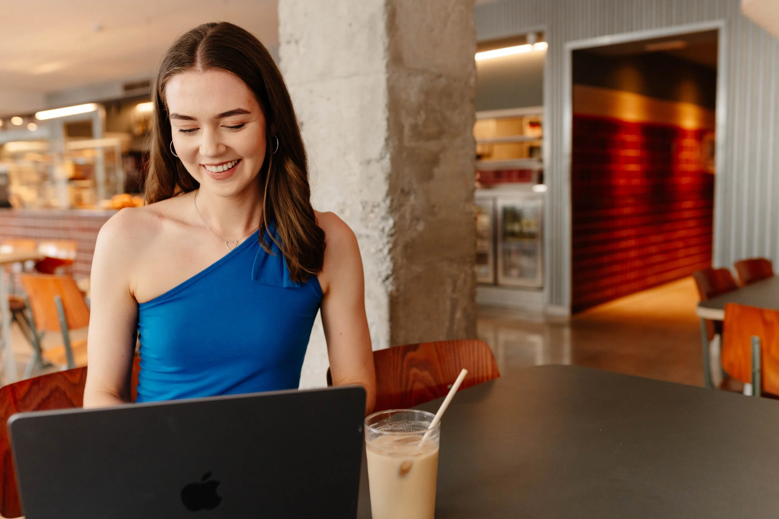 A young woman with long brown hair, wearing a blue one-shoulder top, smiling while looking at a laptop in a modern cafe. There is a glass of iced coffee with a straw on the table.