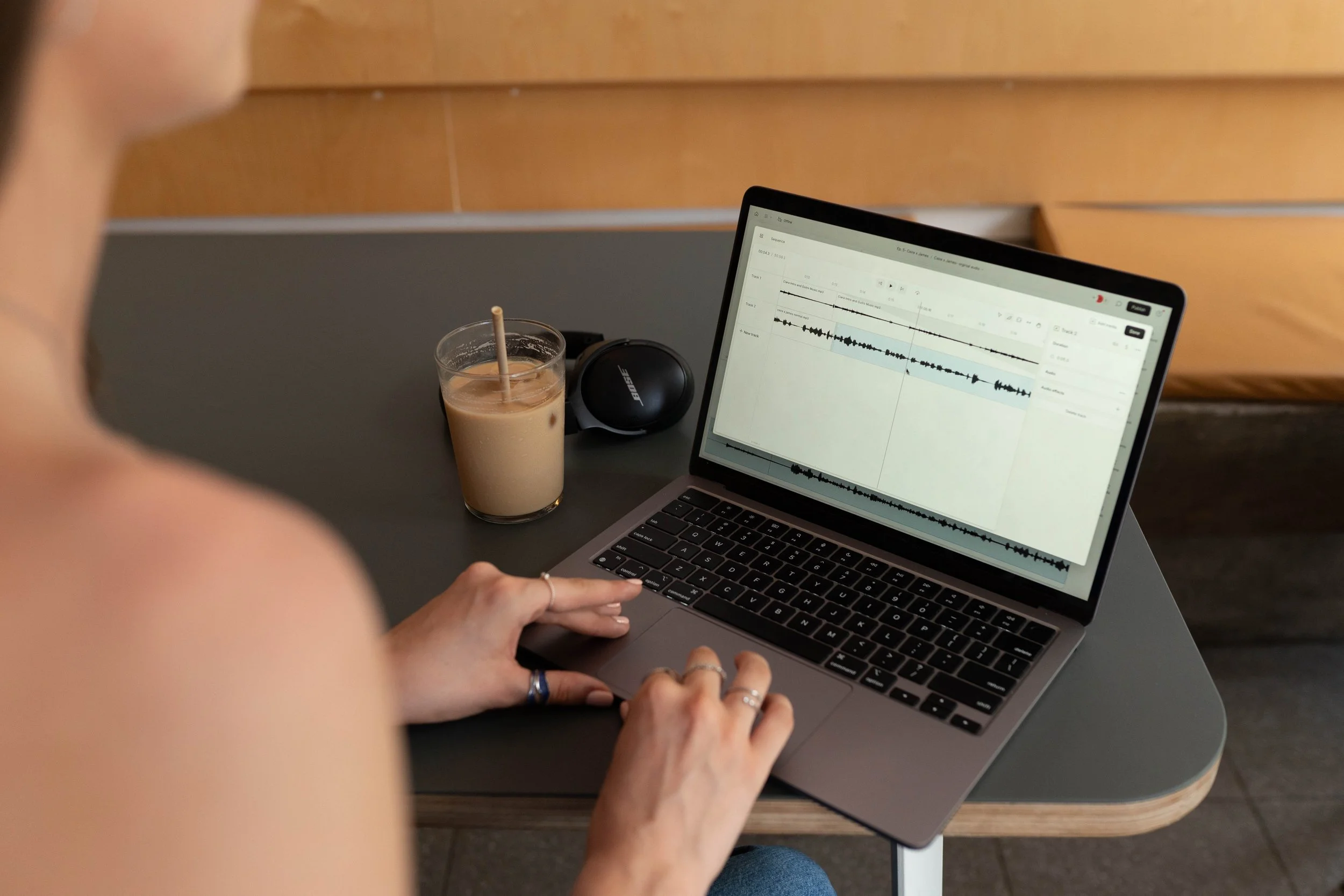 A person working on a laptop with an audio editing software open, a glass of iced coffee with a straw, and a Bose speaker on a dark table.