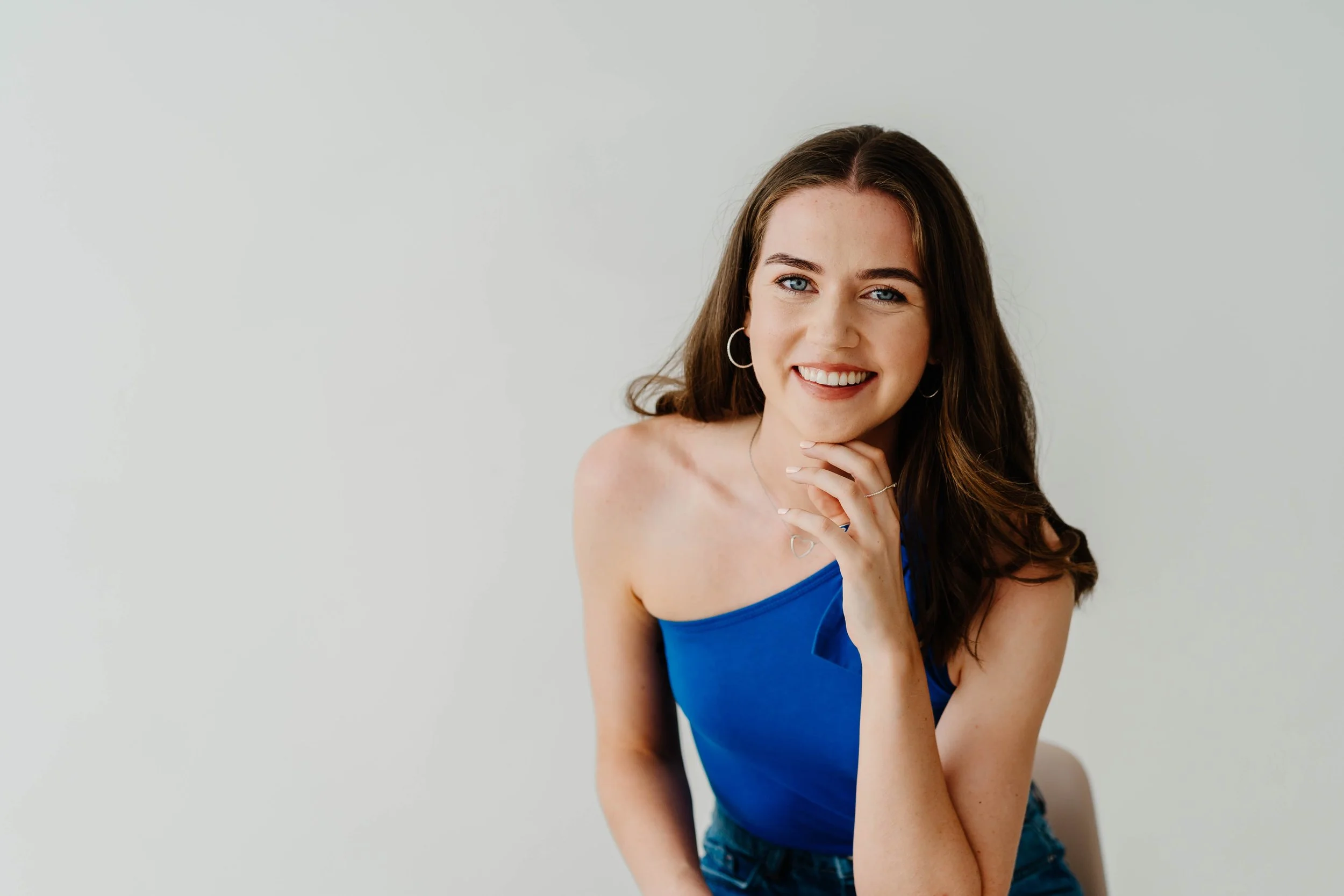 A young woman with long brown hair wearing a blue tank top and silver jewelry, smiling and touching her chin, against a plain white background.