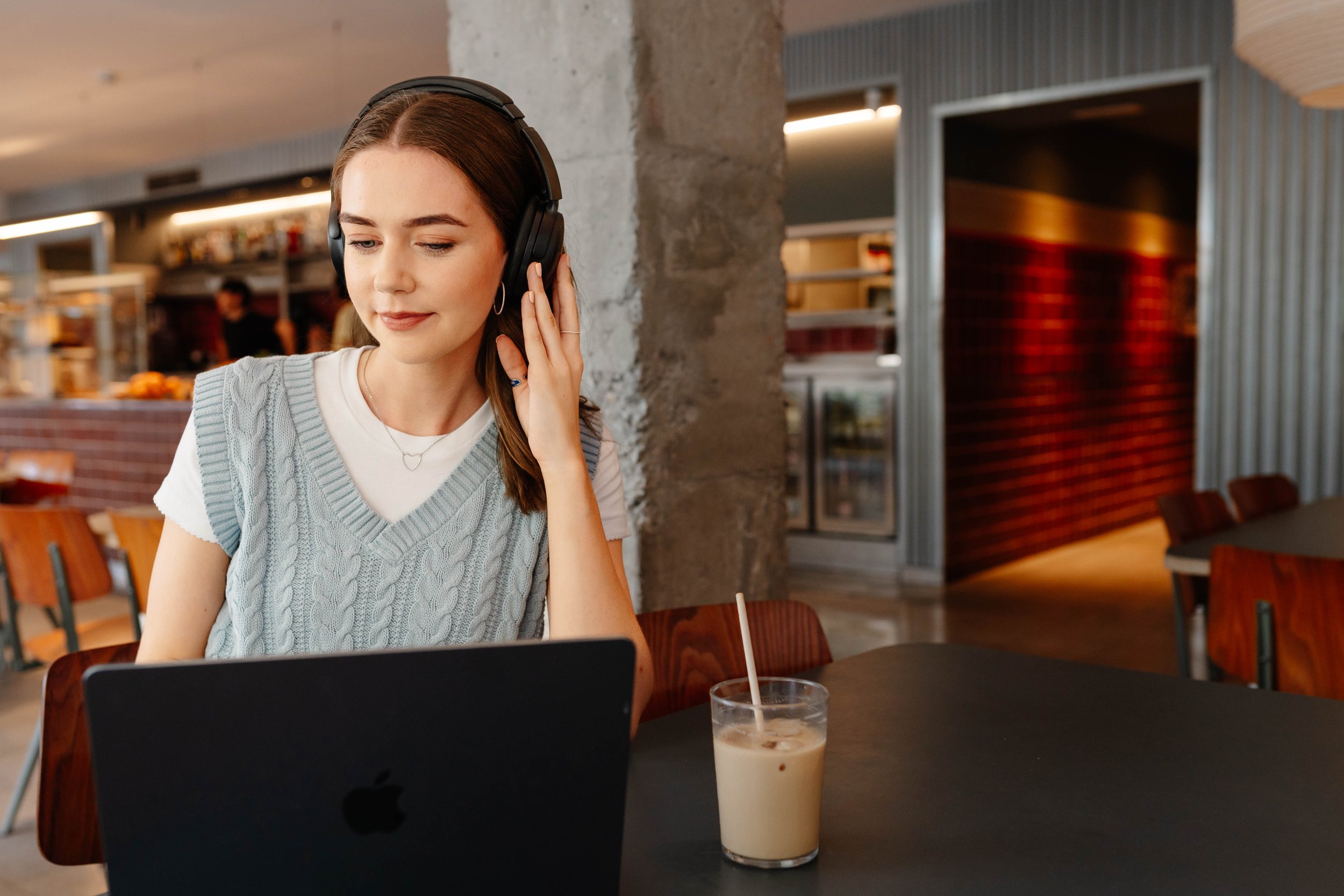 A young woman with brown hair wearing a light blue sweater vest and white t-shirt, sitting at a table in a cafe, wearing large black headphones, looking down at a laptop with a glass of iced coffee on the table.