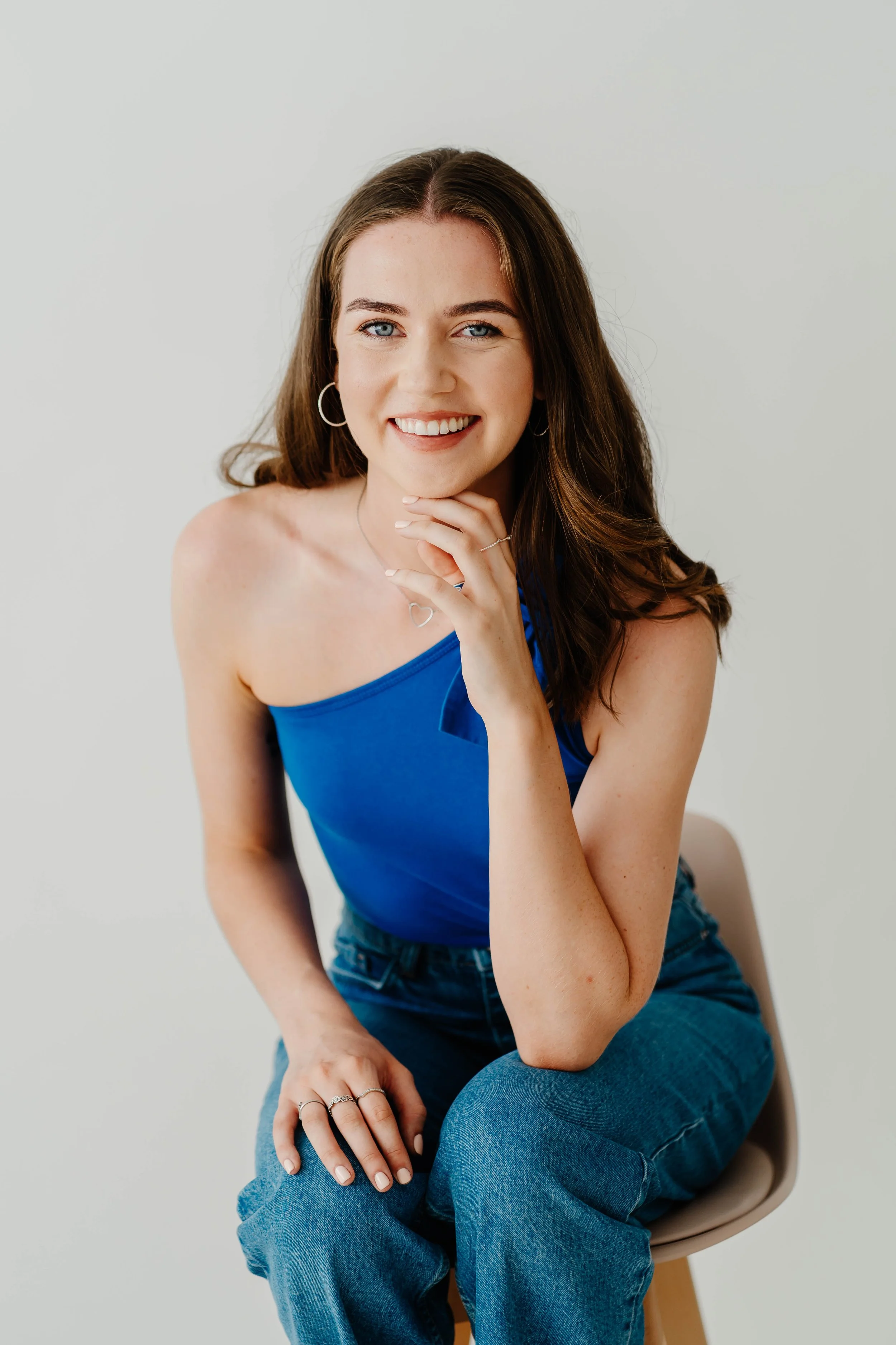 A young woman with long brown hair and blue eyes smiling, sitting on a beige chair against a plain white background, wearing a blue sleeveless top and blue jeans.