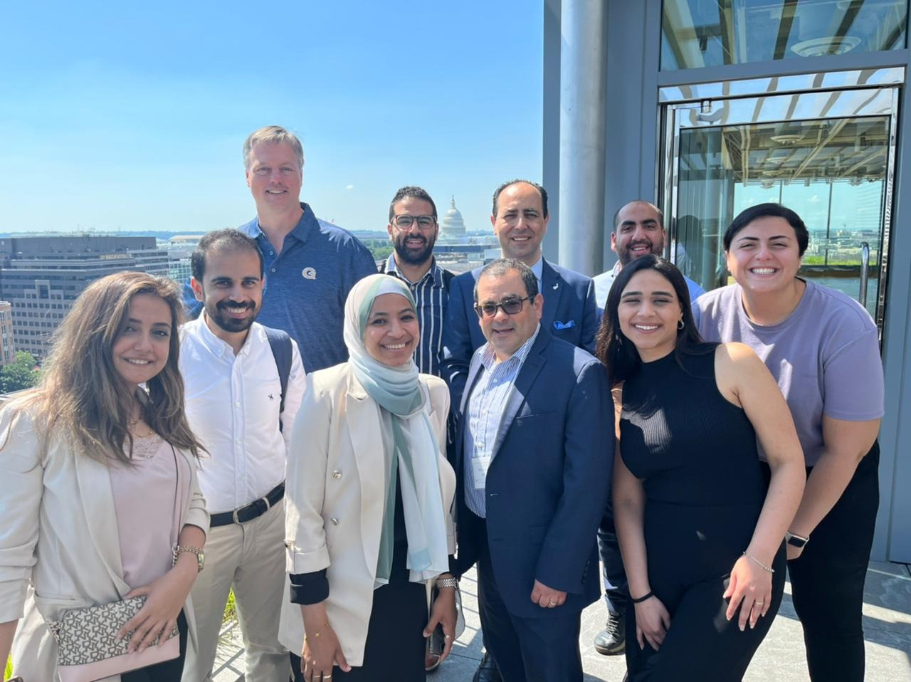 Group of ten people smiling outdoors on a sunny day with city buildings and the US Capitol in the background