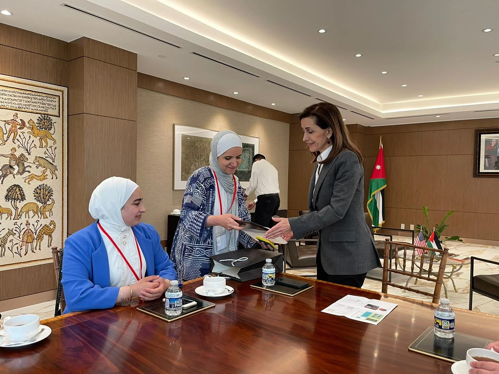 A woman in a gray suit hands a book or document to a woman in a patterned dress, who is standing. A woman in a blue blazer and white hijab is seated at a table, watching. The table has water bottles, cups, and tablets. In the background, a man in a w