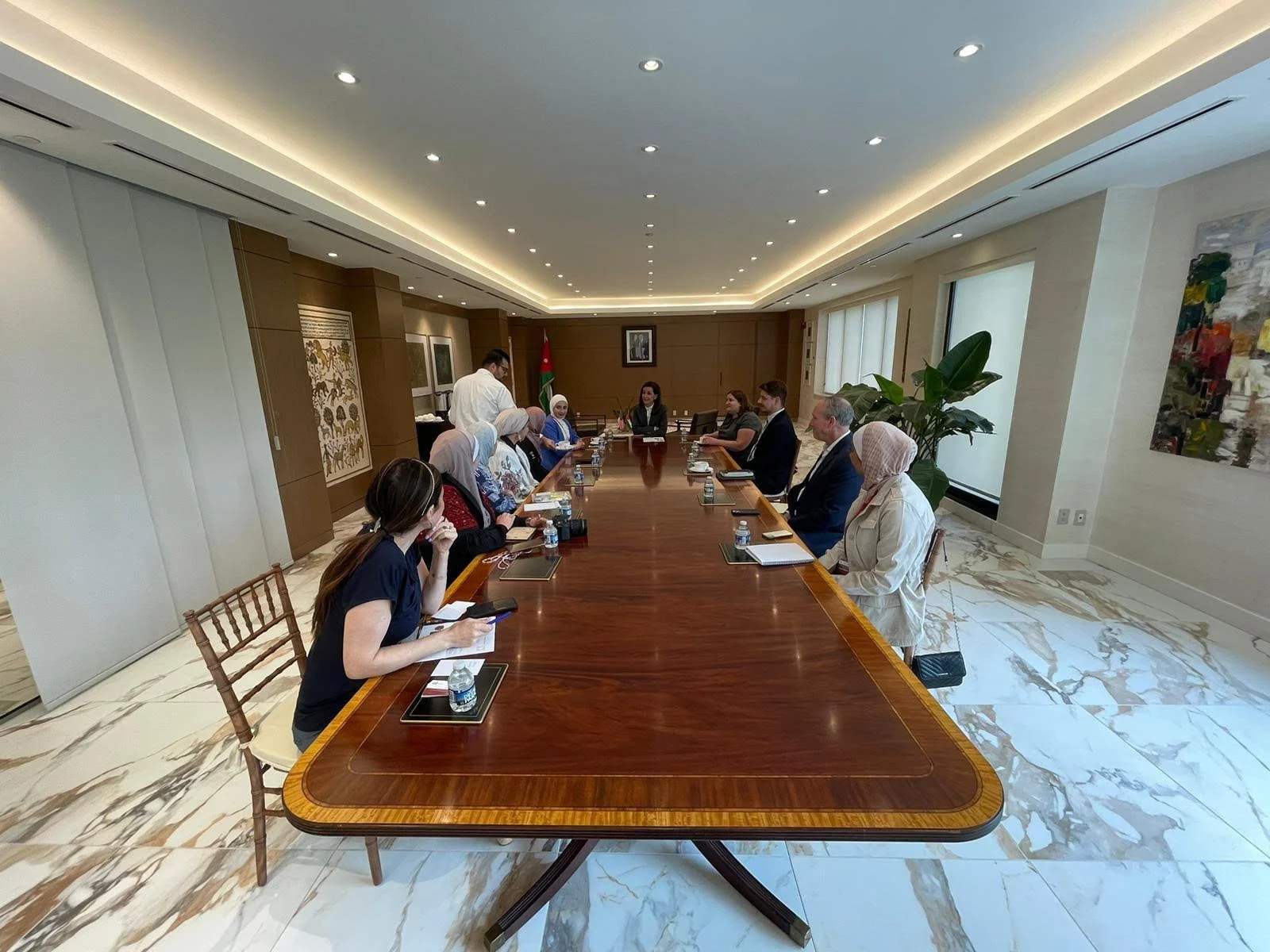 A group of people sitting around a large wooden conference table in a well-lit modern office or meeting room, with water bottles and notebooks in front of them. There are windows on the right side and artwork on the walls.