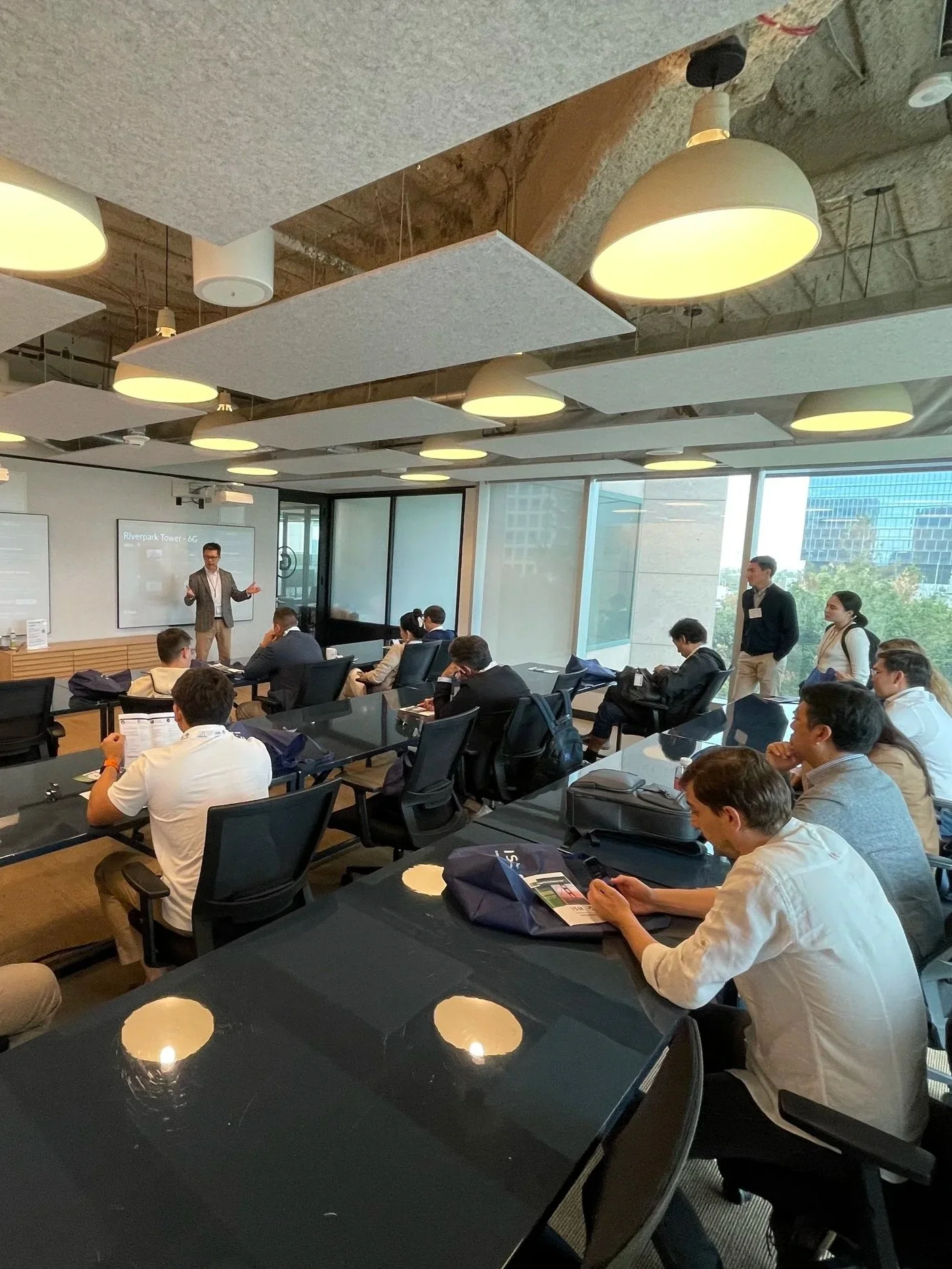 A group of people attending a business presentation in a modern conference room with large windows, a presenter at the front, and people seated at tables listening.