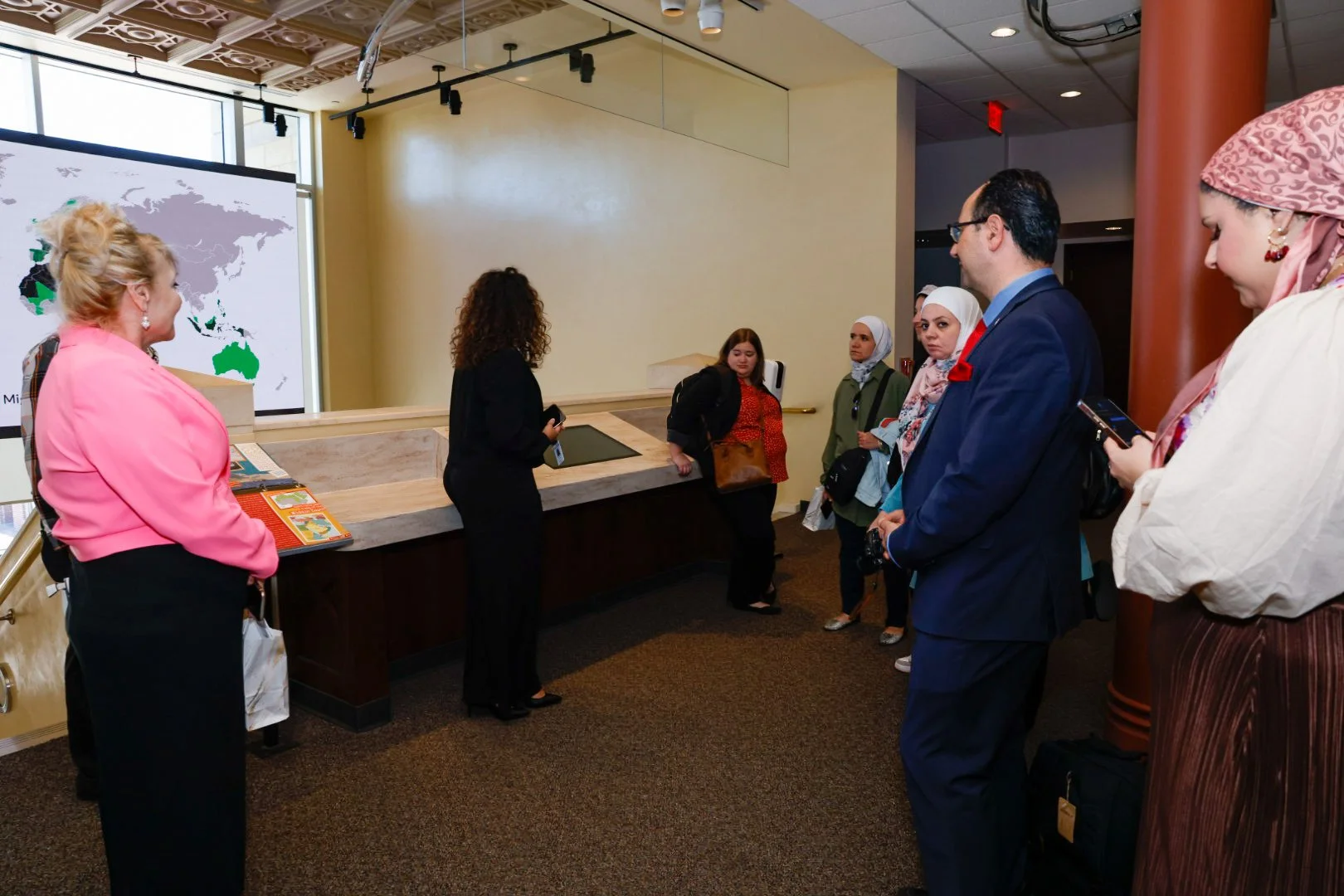 A group of people, including women wearing headscarves, standing in a lobby near the front desk, listening to a woman giving a presentation with a world map projection on a large screen in the background.