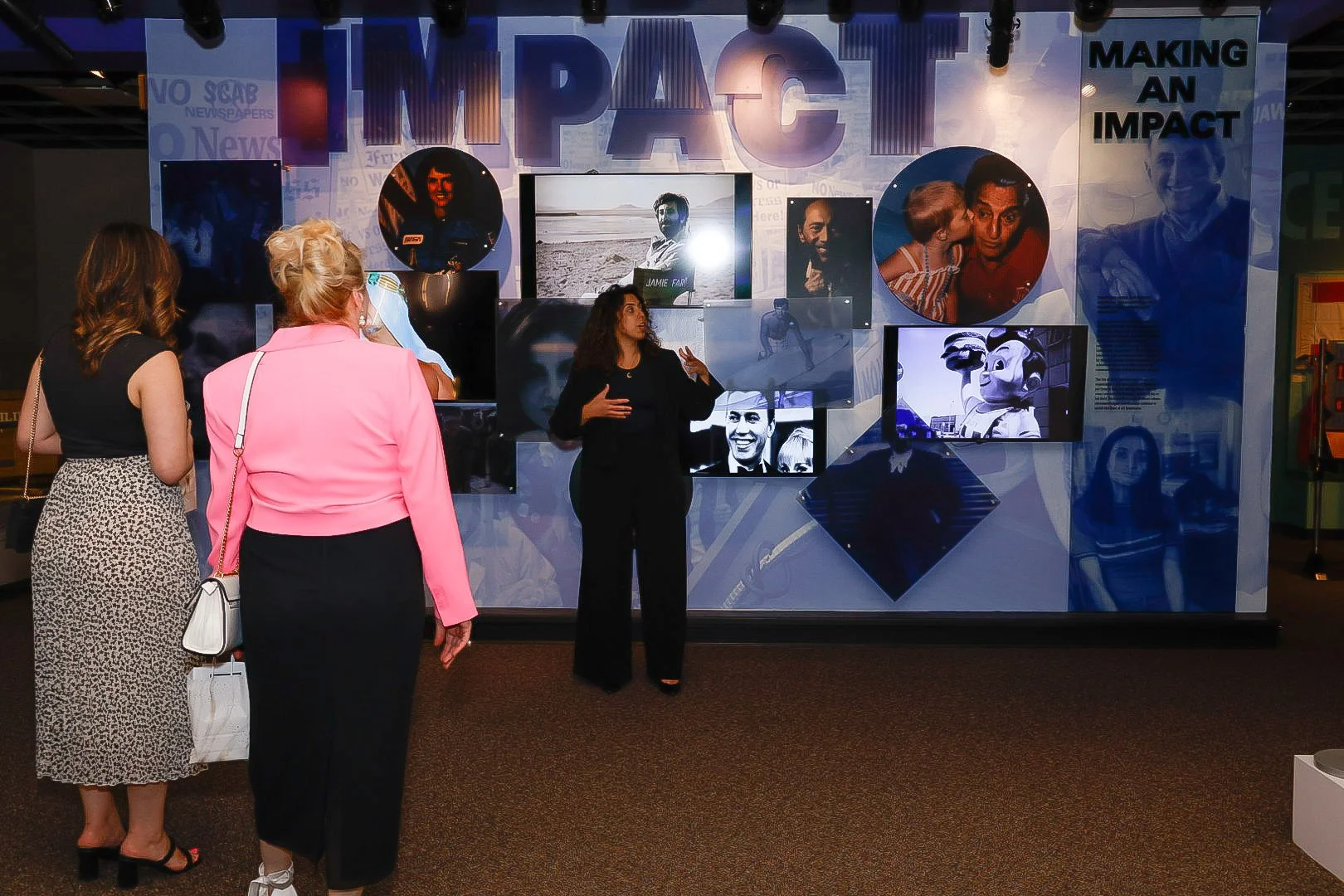A woman explains a display titled 'IMPACT' to two women at an indoor event, with the display featuring photos and the words 'MAKING AN IMPACT'.