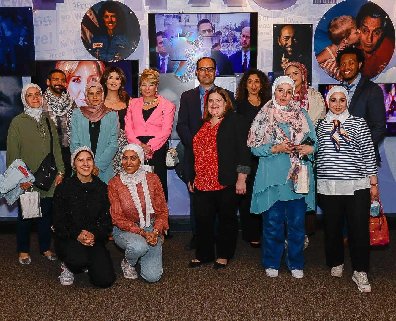 Group of diverse people posing for a photo in front of TV screens with images of astronauts and TV personalities in a studio or event setting.