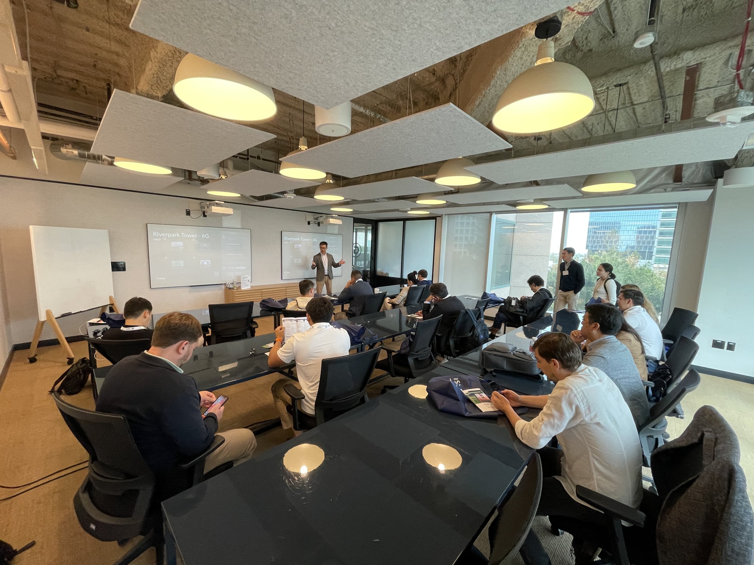 A group of people attending a business presentation in a conference room, with a presenter standing at the front near two screens displaying slides titled 'Riverpark Tower - 6G'. Some attendees are looking at their phones, while others are listening 