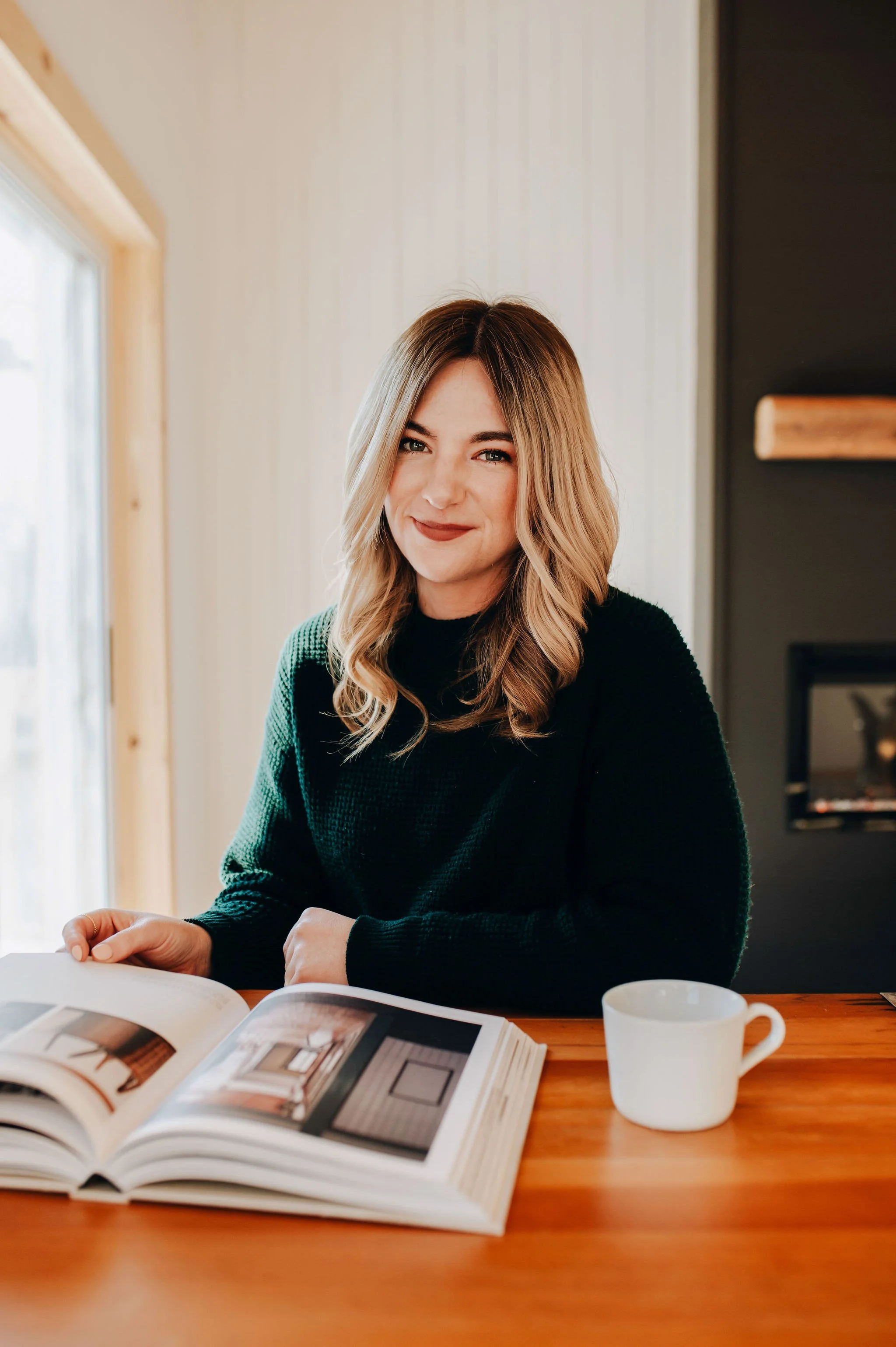 A woman with wavy blonde hair and a black sweater sitting at a wooden table, smiling slightly, with an open magazine and a white mug in front of her, near a window in a cozy room.