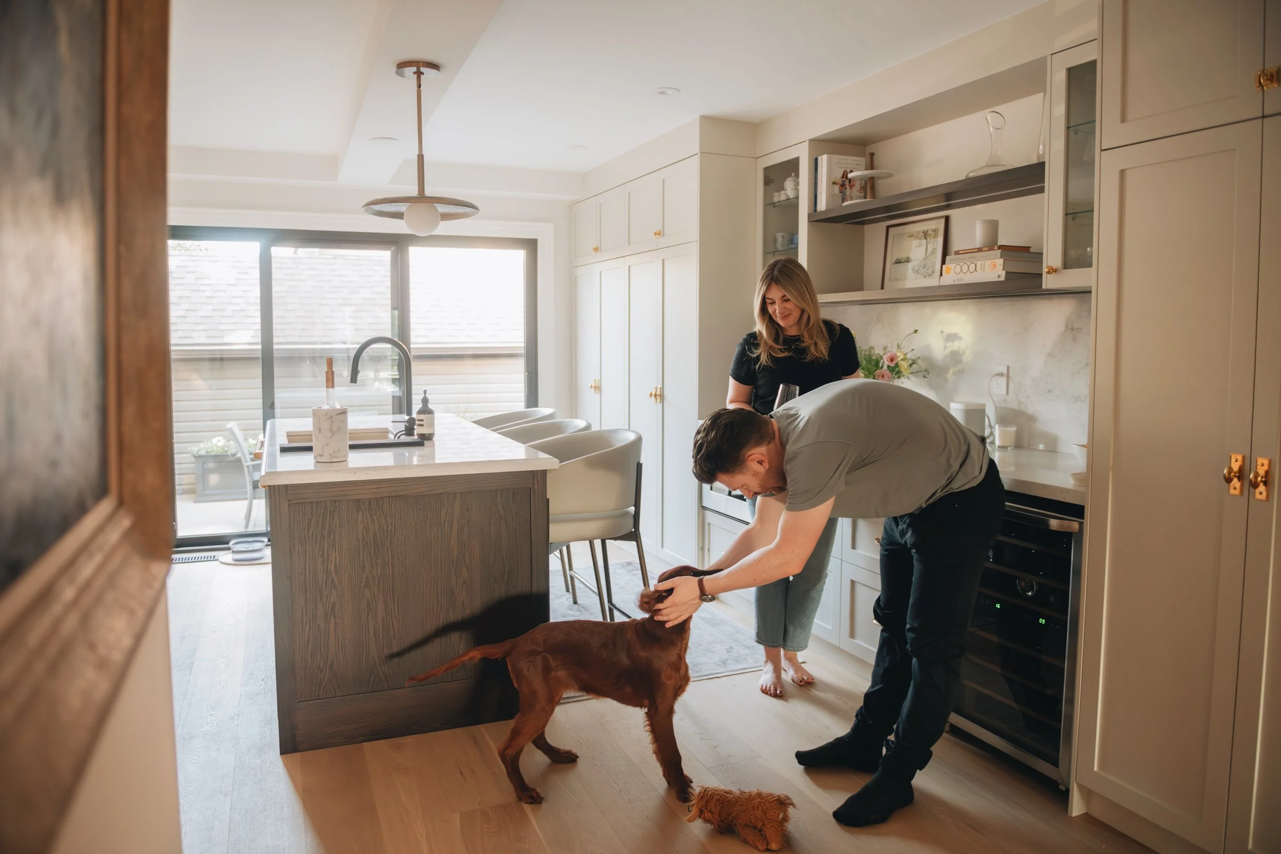 A couple playing with their dog in a bright kitchen. The man is bending down and petting the dog, while the woman watches and smiles. The kitchen has white cabinets, a marble backsplash, and a large window letting in natural light.