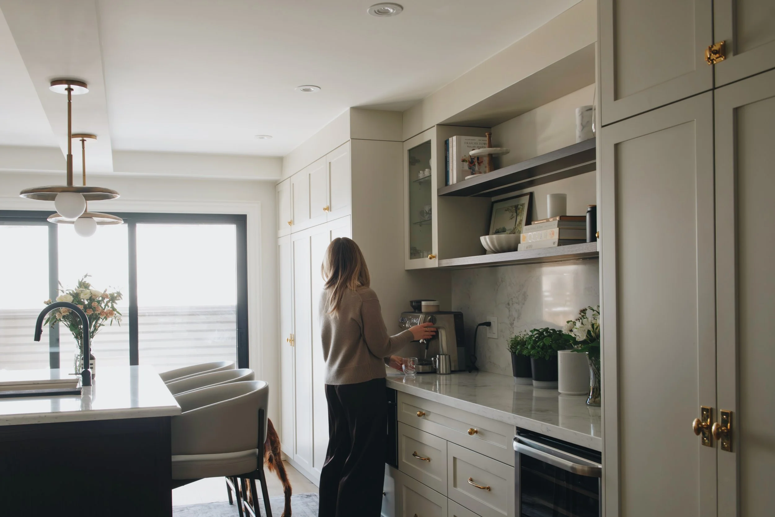 A woman in a beige sweater is making coffee in a modern kitchen with white cabinets, a marble countertop, and open shelves with books and decorative items. There is a large window with natural light and a dining table with flowers nearby.