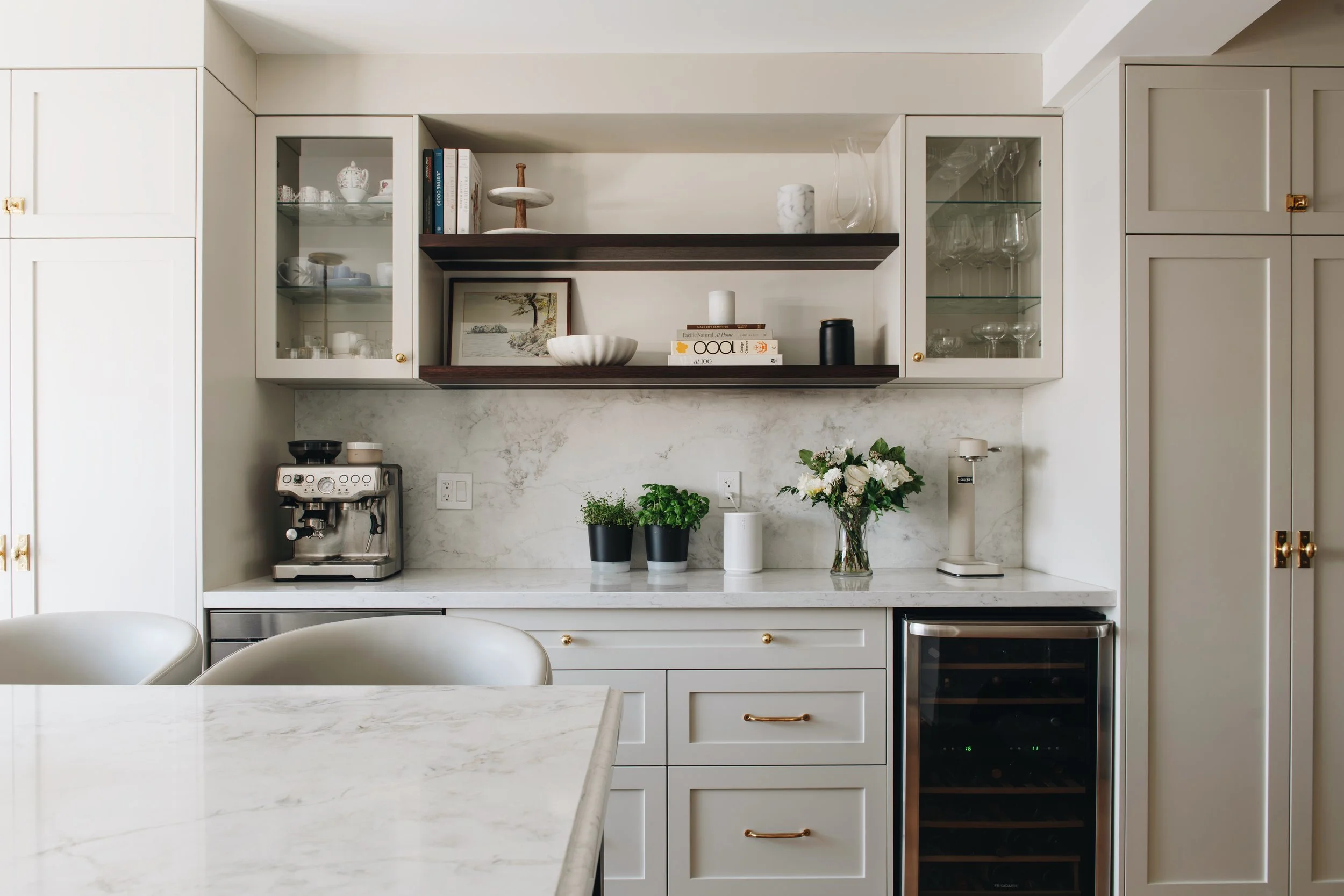 Modern white kitchen with marble countertops, open shelving with decorative items, glassware, a coffee machine, potted plants, and a built-in wine cooler.