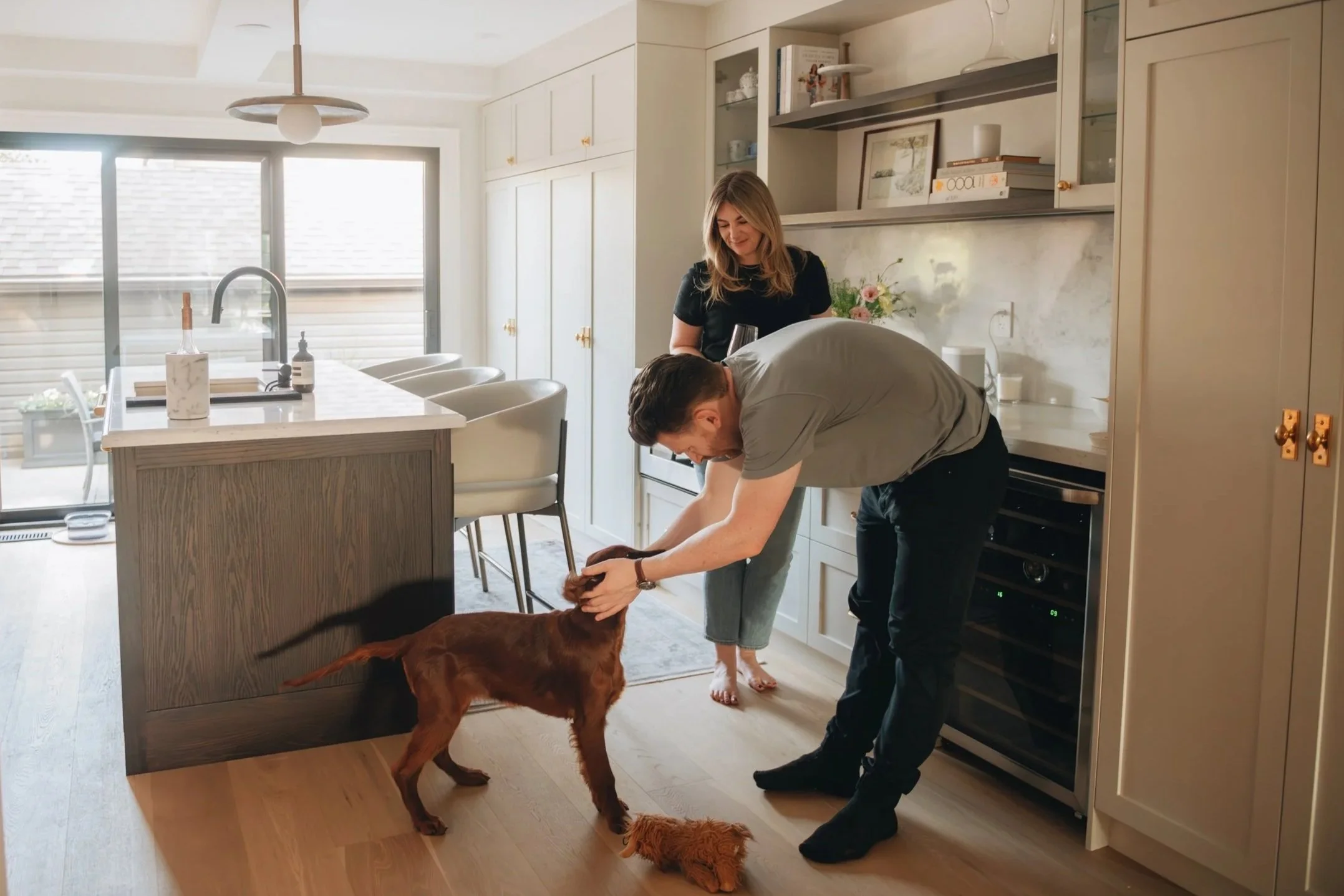 A woman and a man are in a kitchen with a dog. The woman is smiling and looking down at the man, who is bending over and petting the dog. The dog is a brown Labrador Retriever, and there is a small plush toy on the floor nearby. The kitchen has white cabinets, a marble countertop, and a dining area with a table and chairs. Large windows let in natural light.