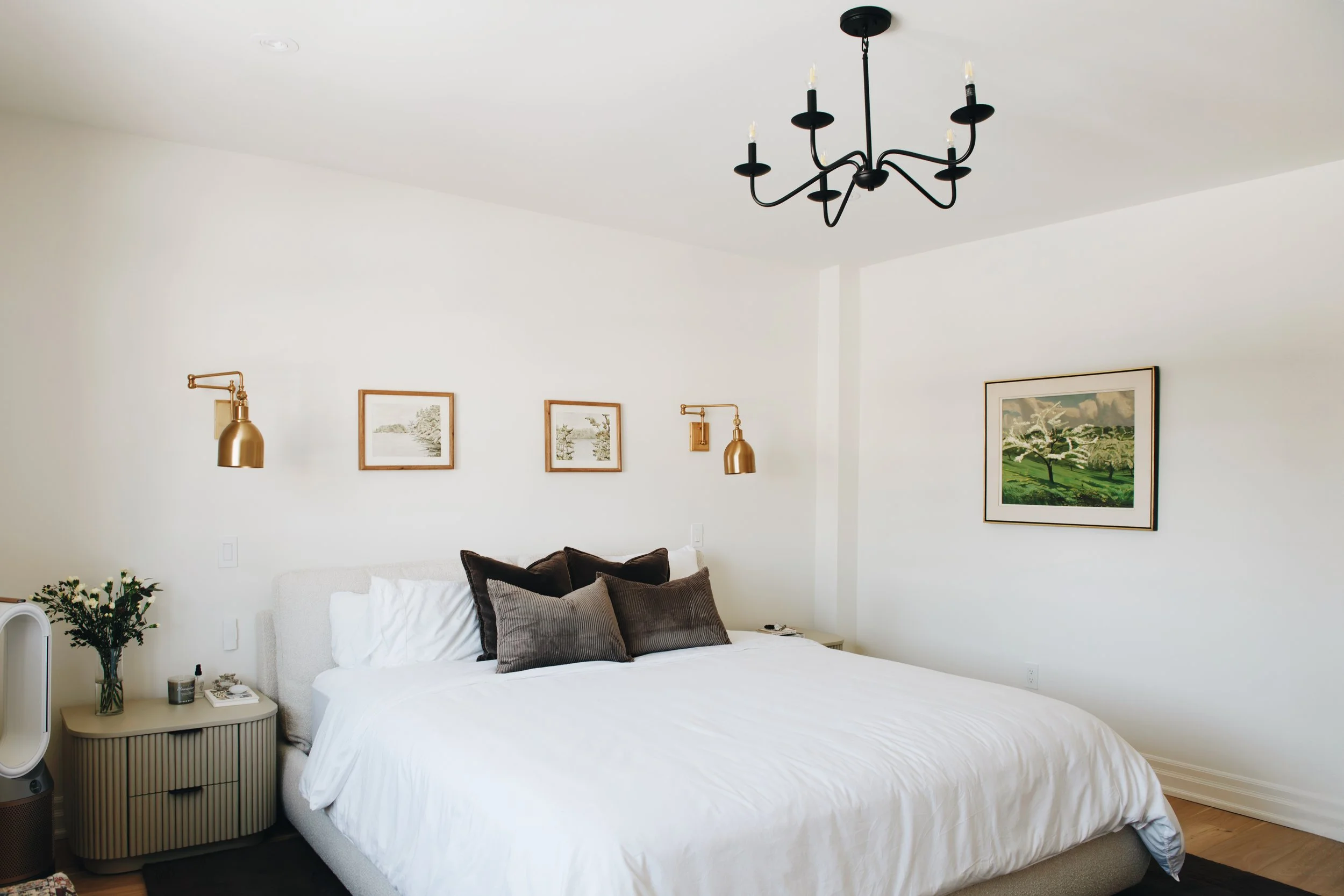 A bedroom with a white bed, brown and gray pillows, and framed artwork on the white wall. A black chandelier hangs from the ceiling, and gold wall sconces are mounted on the wall above the bed. A small nightstand with decorative items is near the bed.