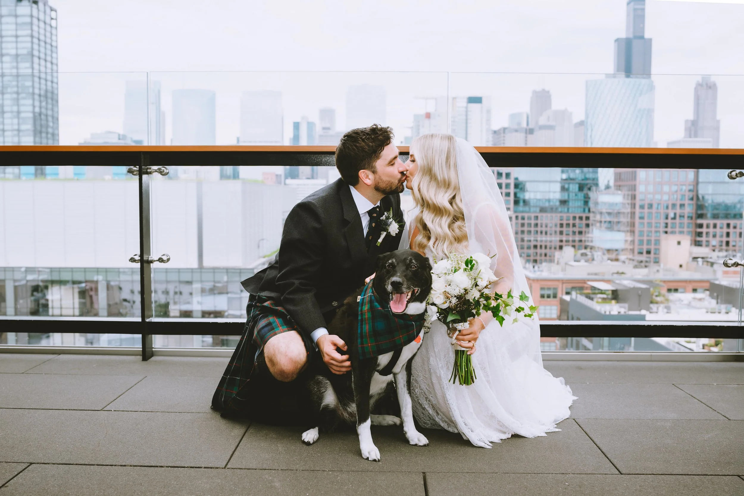 Bride and Groom kissing while posing with their dog in front Chicago skyline on their wedding day.