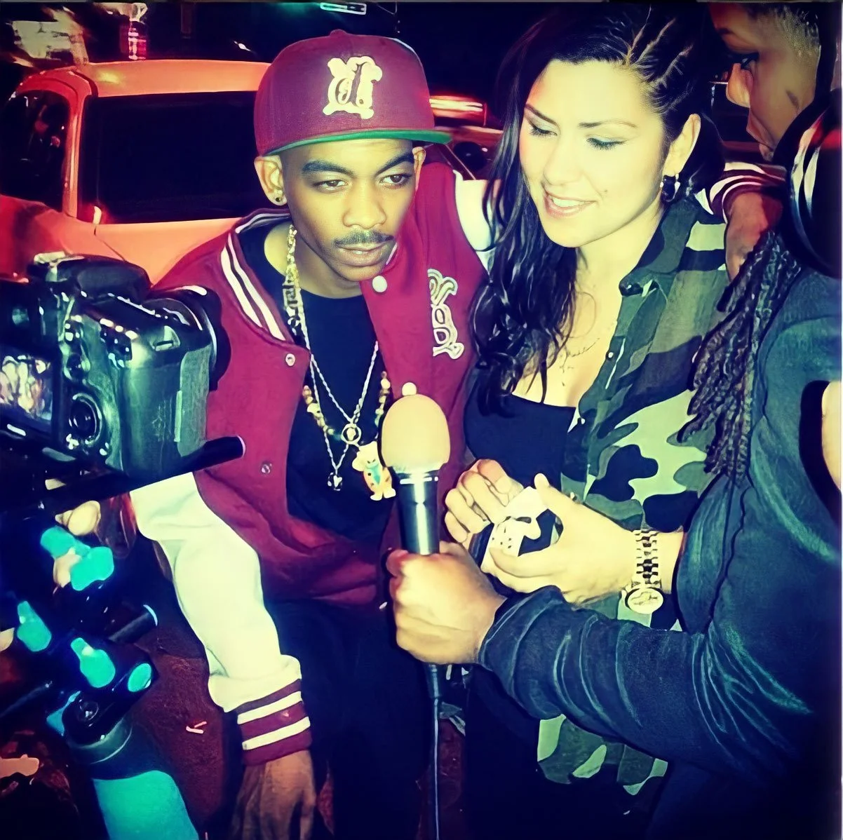 A man in a maroon baseball cap, maroon and white jacket, and chains takes a selfie with two women holding a microphone and a smartphone, in front of a red car at night.