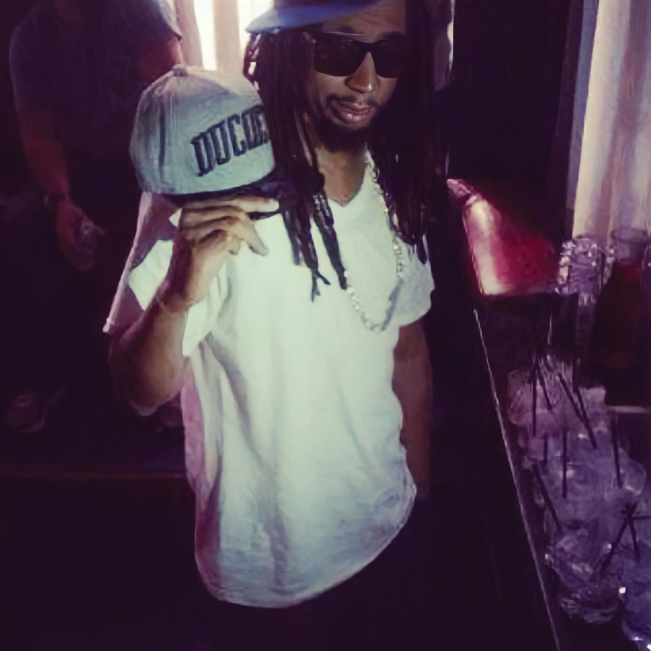 A man with dreadlocks wearing sunglasses and a white t-shirt poses with a young child on his shoulder, who is wearing a gray cap with the word 'DUCARS' on it, in an indoor setting with other people and chairs in the background.