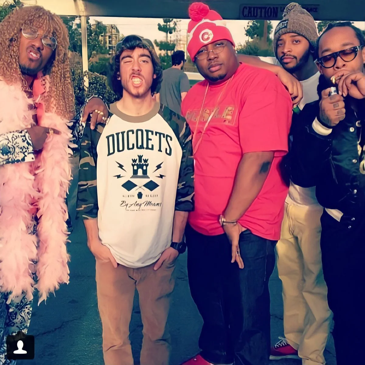 Group of five young men standing outdoors, dressed in casual and trendy clothing, some wearing hats and sunglasses, posing for the photo.